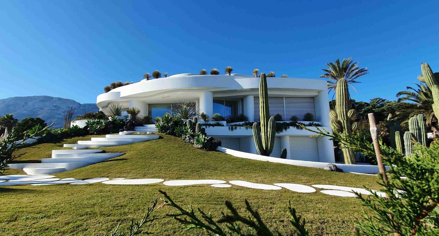Modern white curved house on a grassy hill with cacti and palm trees against a mountain backdrop and clear blue sky.