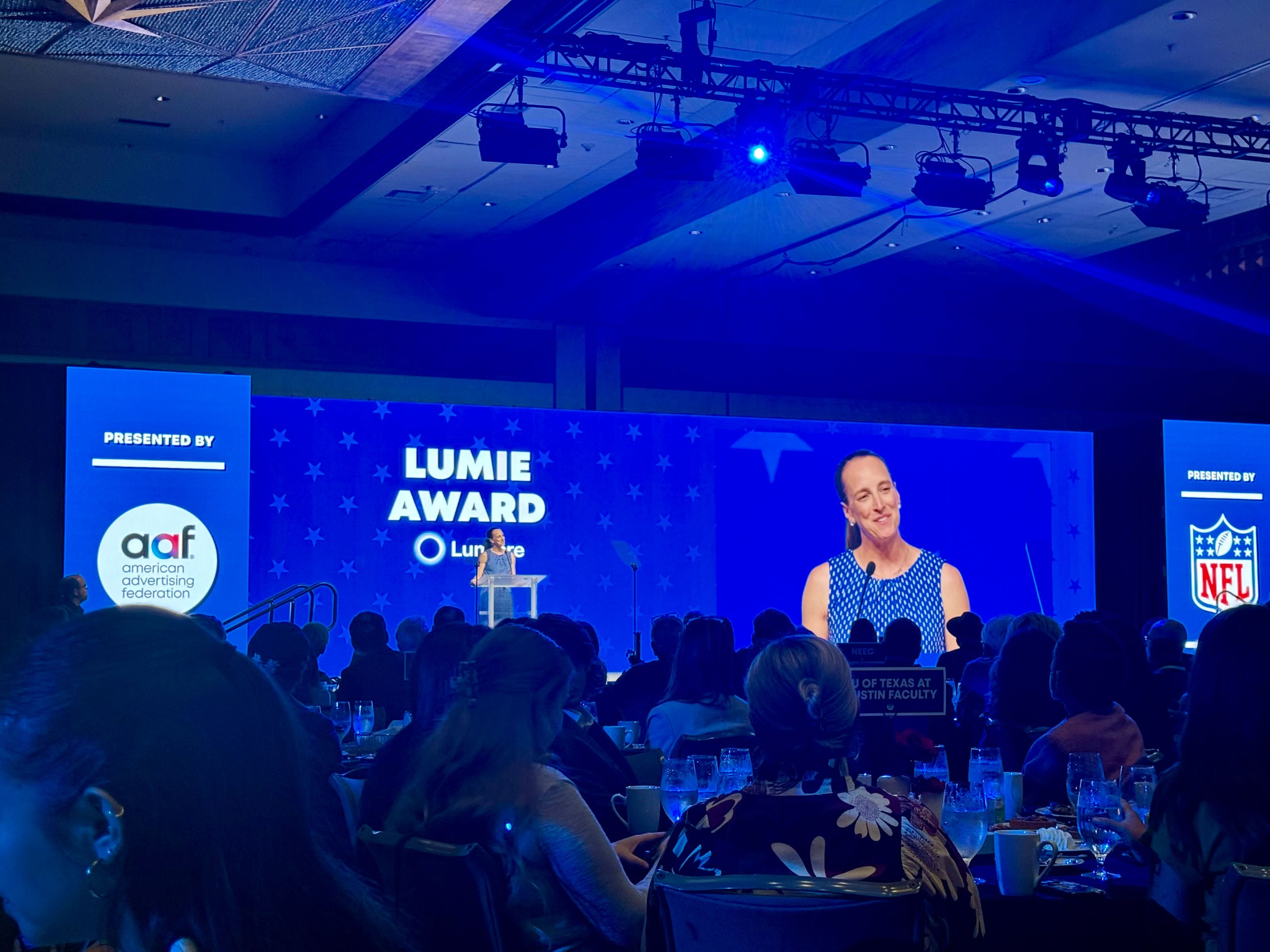 A blue-tinted photo of an indoor awards ceremony. The white Latitude logo appears on the left and the American Advertising Federation (AAF) logo on the right. In the background, a speaker stands at a podium on stage beside large screens reading 'Lumie Award' and showing a close-up of the presenter, Kimberly Kelling . A silhouetted audience fills the foreground