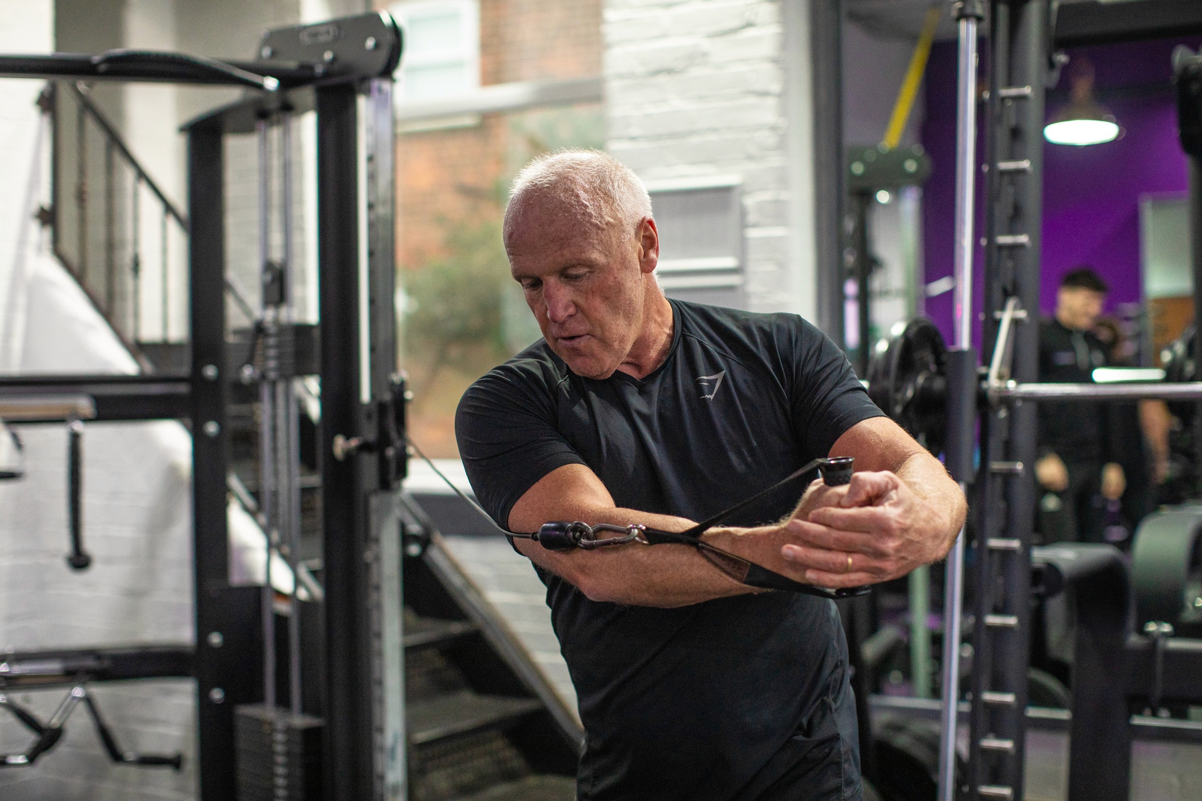 A man in athletic wear uses a cable machine in a gym, performing a cable crossover exercise. The room has gym equipment and a focused atmosphere.