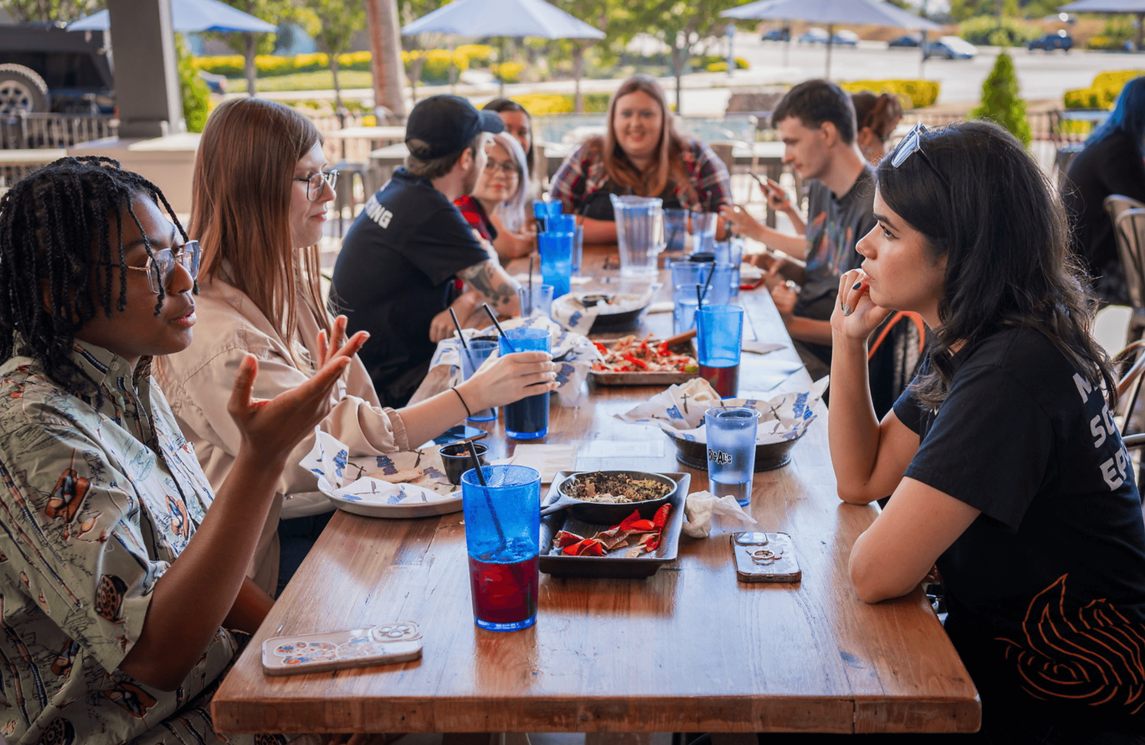 A group of people is engaged in lively conversation while sitting around a wooden table at an outdoor restaurant, featuring blue water glasses and dishes of food.