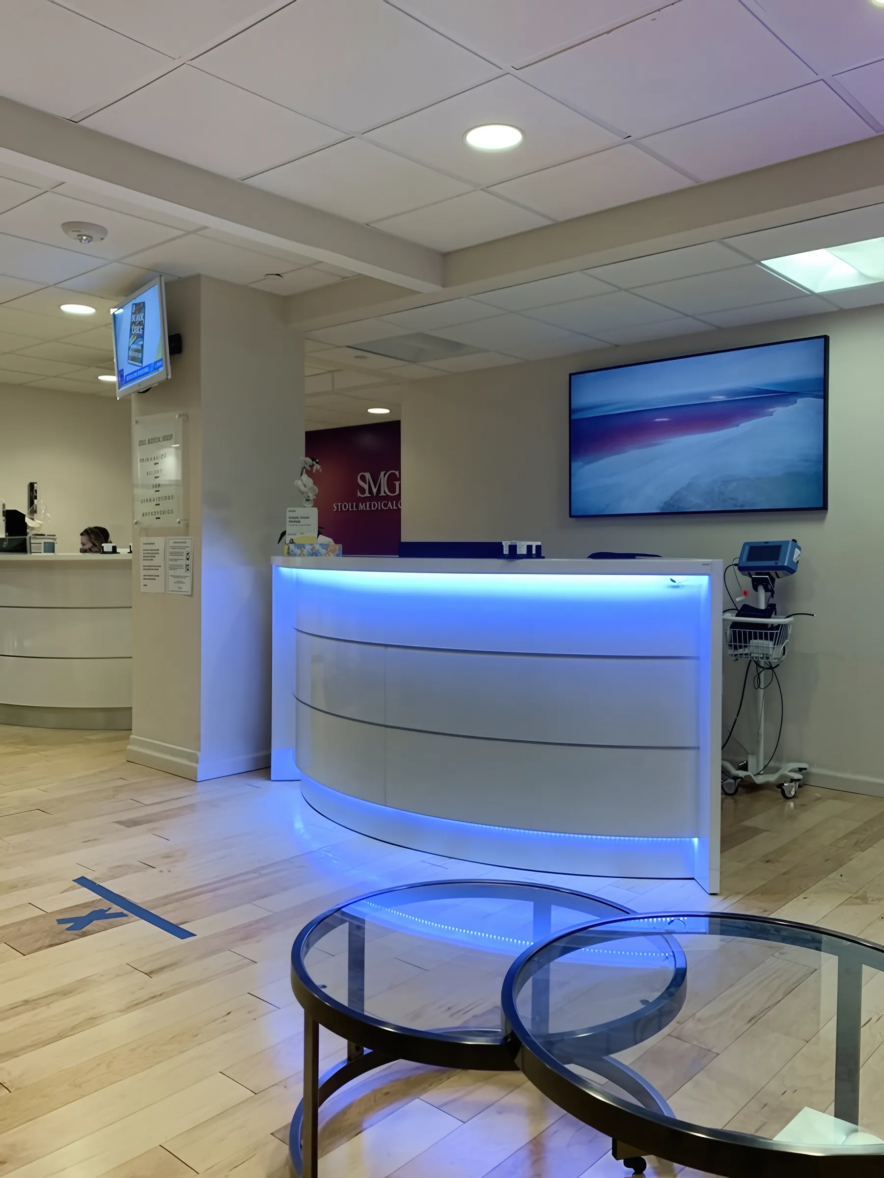 Hardwood flooring in a Stoll Medical Group reception area featuring an LED-lit curved white front desk, wall-mounted TV, and medical equipment.