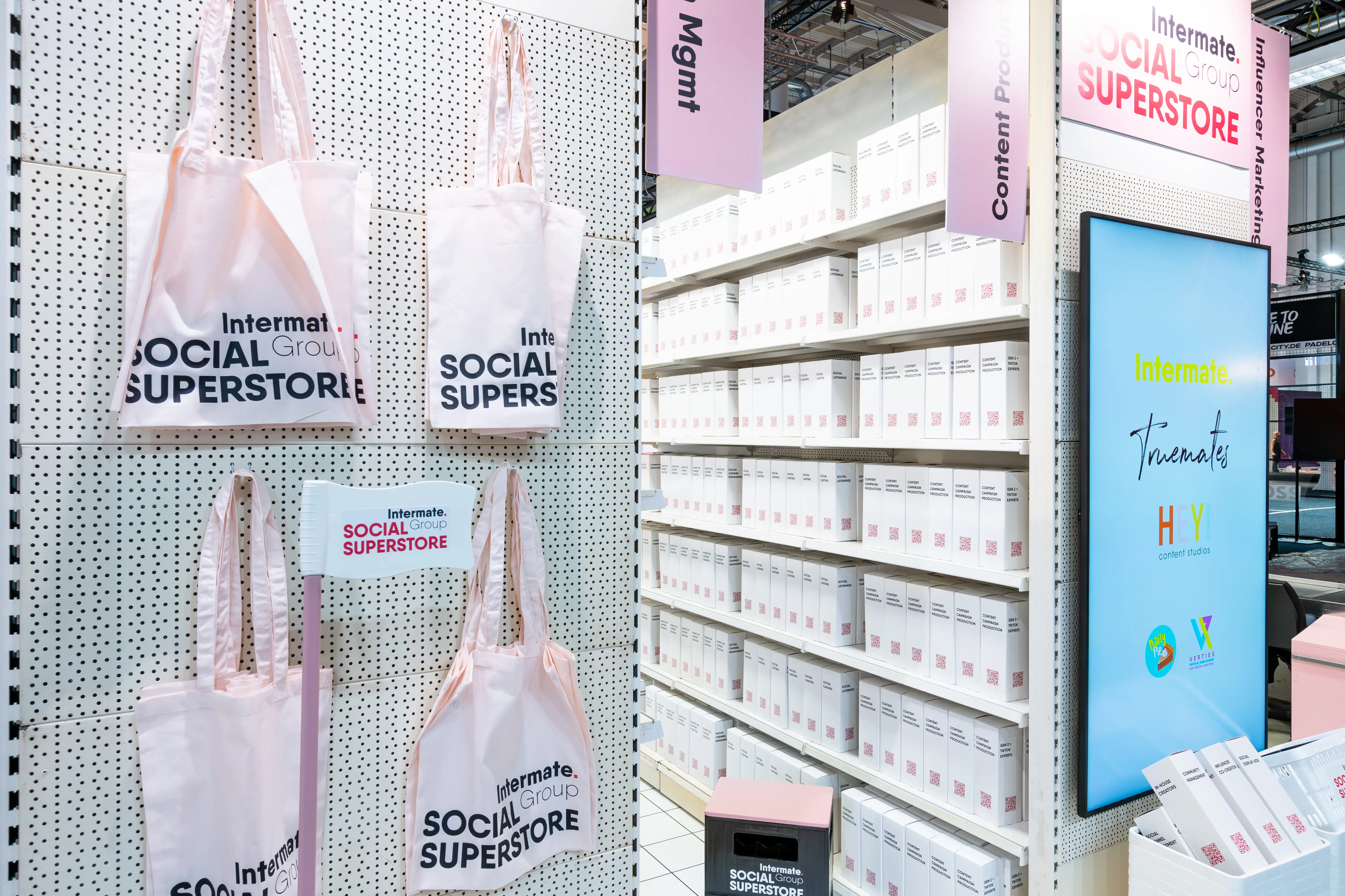Pink Social Superstore tote bags on a perforated wall next to shelves of white product boxes at the Intermate booth.