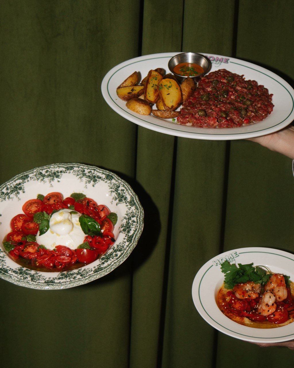 A vibrant table setting featuring a plate with seasoned steak tartare and roasted potatoes, a dish of sliced tomatoes and mozzarella garnished with basil, and a plate of spicy shrimp placed in a rich sauce, all against a dark green curtain backdrop.
