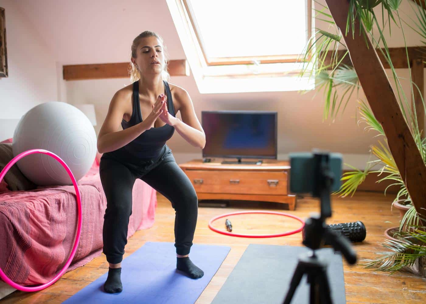A woman in bedroom doing a workout guide while looking at her phone on a tripod in front of her
