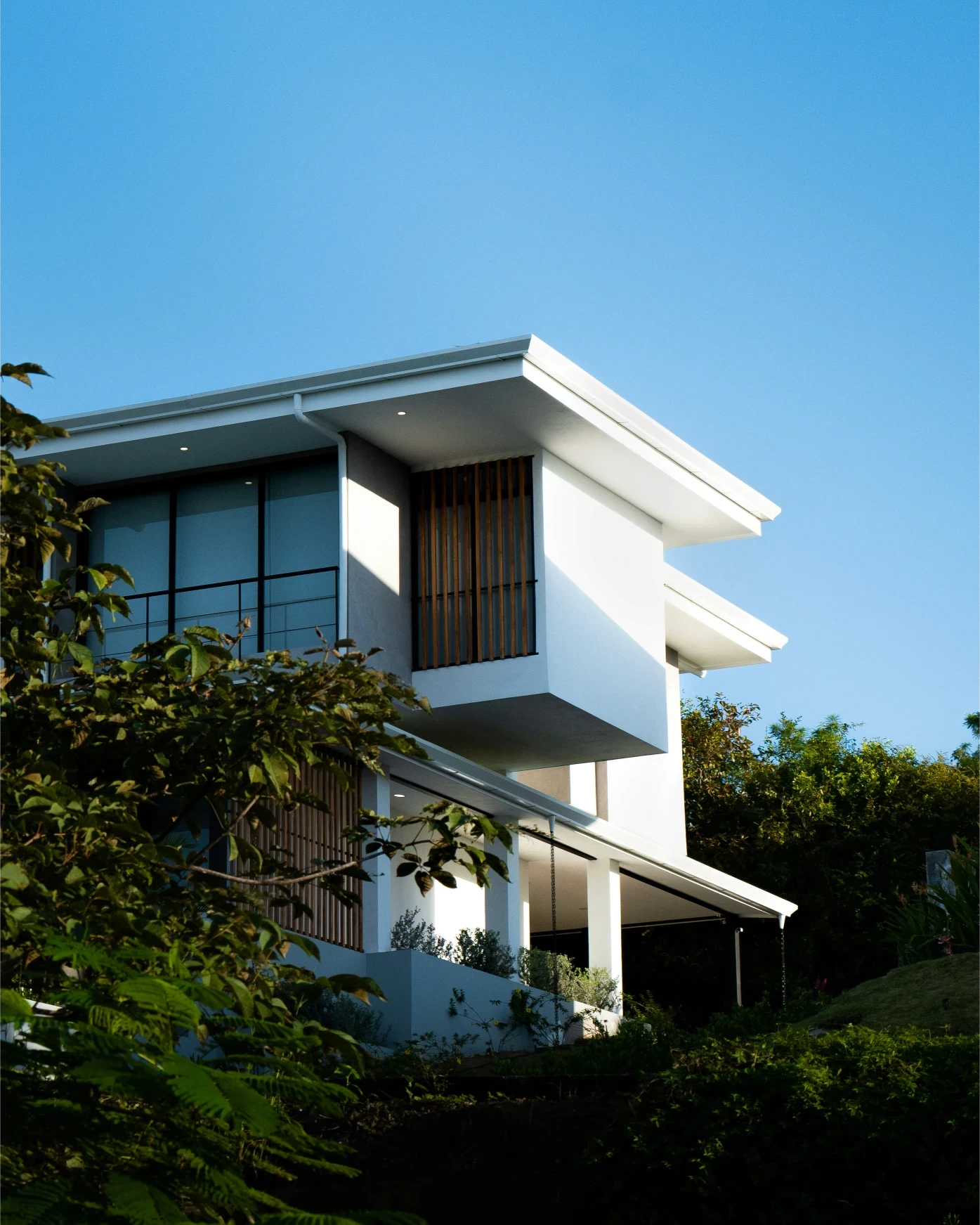 A modern house with large windows and a flat roof, surrounded by greenery against a clear blue sky.