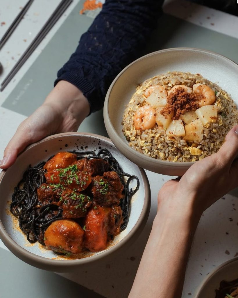 Two hands holding bowls of Taiwanese dishes, featuring black noodle dish with chicken and a plate of fried rice topped with prawns and garnished with herbs.