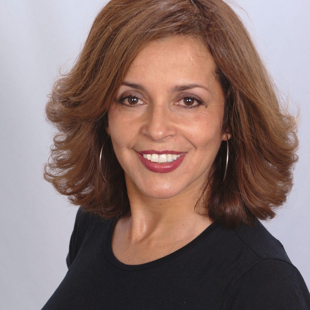A young person with curly hair smiles at the camera while sitting in front of a laptop against a dark background.