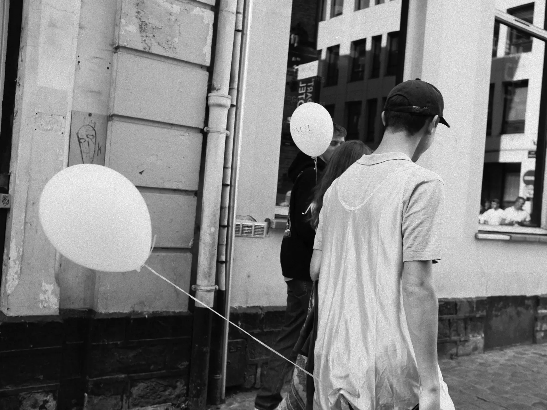 Man walking beside a white balloon on a narrow street, in black and white