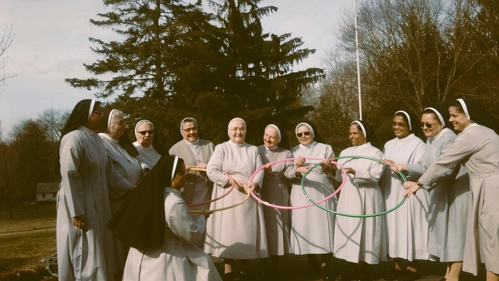 Group of twelve Catholic nuns from the Daughters of Our Lady of the Garden, USA province, standing outdoors in a semicircle. Dressed in light gray habits with black veils, the sisters hold colorful hula hoops in a joyful, recreational moment. Set against a winter landscape with leafless trees and an evergreen, this image reflects the congregation’s charism of Vigilant Evangelical Charity and their commitment to joyful service and community life.