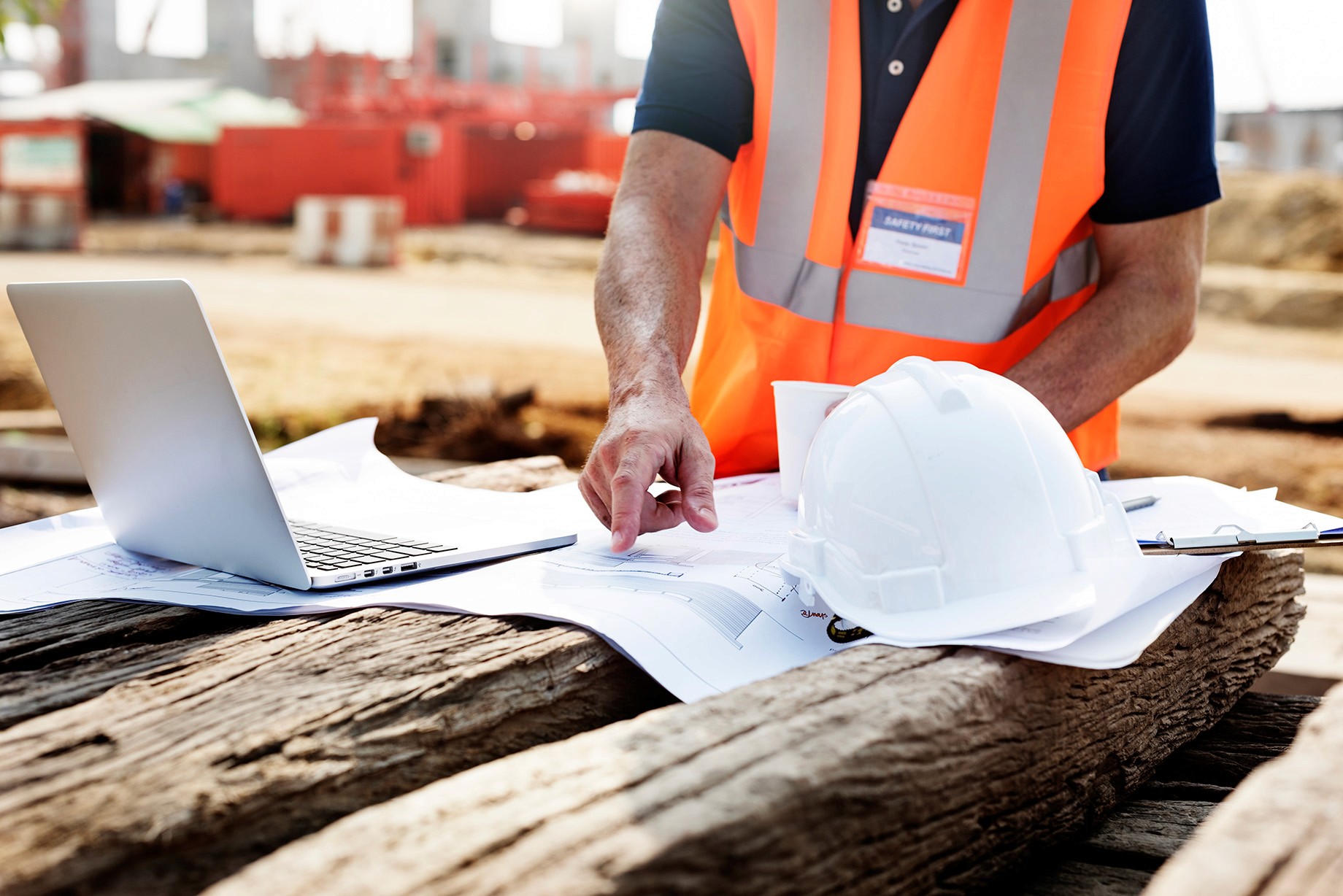 Construction worker in a high-visibility vest reviews blueprints with a laptop and hard hat on a wooden surface.