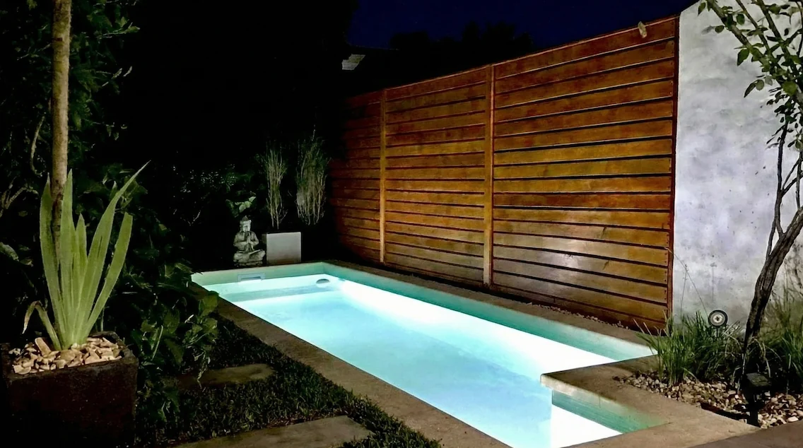 Pileta de diseño angosto optimizada para patios, con iluminación subacuática, cerco de madera rústico y decoración zen con estatua de Buda.