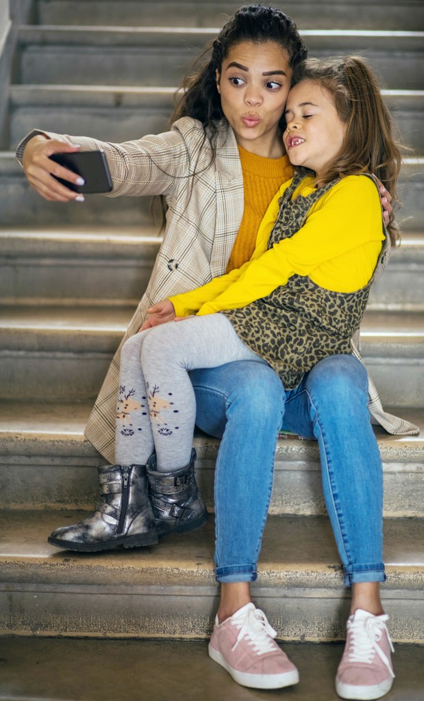 The image captures a heartwarming moment between two sisters sitting on a staircase. The older sister is taking a selfie with a playful pout, while the younger sister, dressed in a yellow sweater and leopard-print dress, leans in with a big smile. Their casual attire and the concrete steps create a relaxed and intimate atmosphere, highlighting their close bond and joyful interaction.