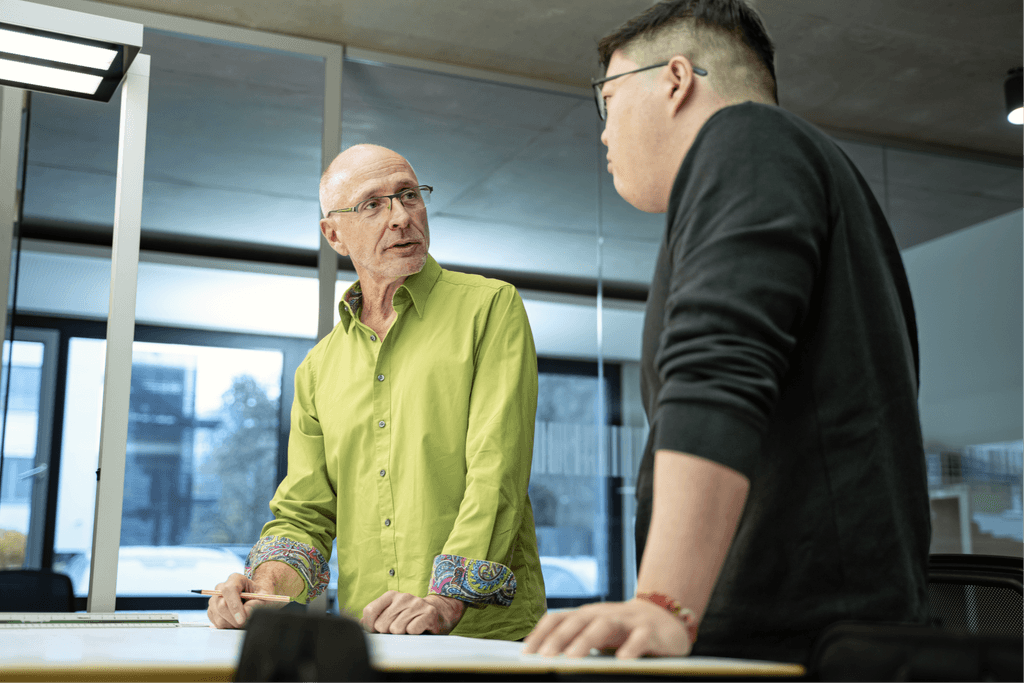 Zwei Mitarbeitende der kba Architekten und Ingenieure GmbH stehen an einem großen Planungstisch und diskutieren technische Bauzeichnungen; der ältere Mann im grünen Hemd erklärt fachliche Details, während sein Kollege aufmerksam zuhört.