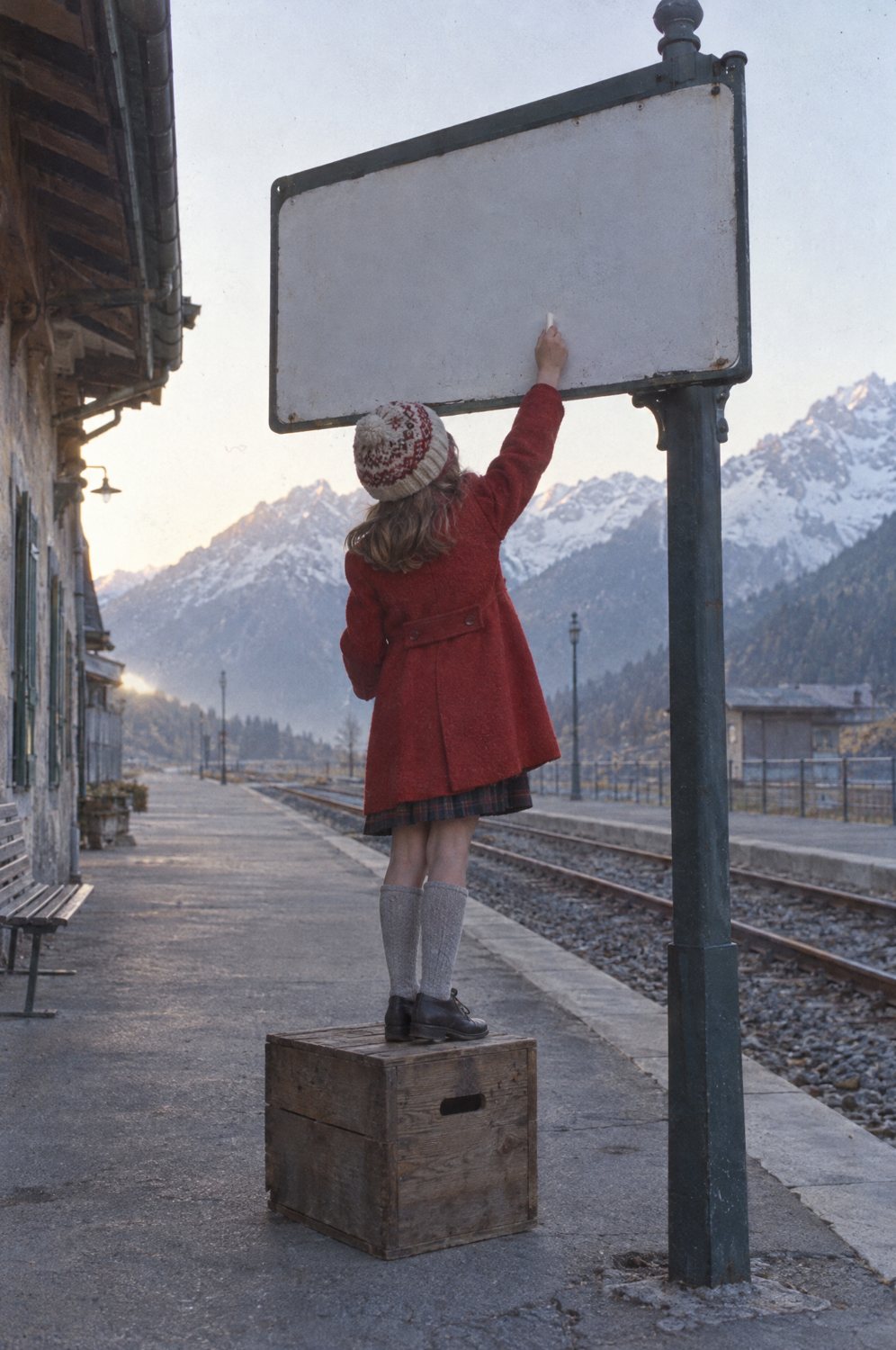 A girl in a red coat stands on a crate and reaches up with chalk to the blank station sign