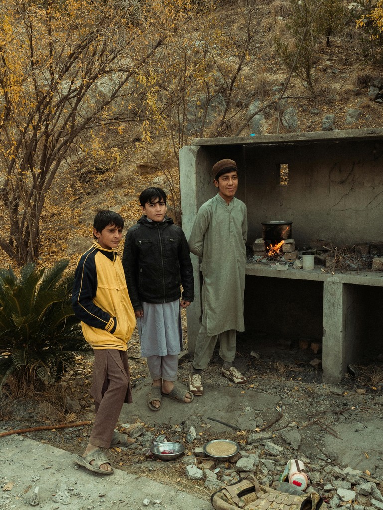 Three boys in traditional attire stand near an outdoor cooking area with a fire, set against a backdrop of autumnal trees and rocky terrain, illustrating daily life in a rural setting.