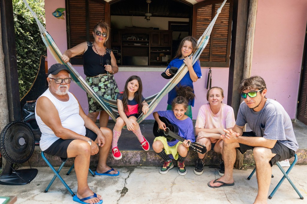 A multi-generational family poses happily in front of their home, with the adults seated and the children sitting on a hammock and holding musical instruments, creating a joyful and relaxed atmosphere in an outdoor setting.