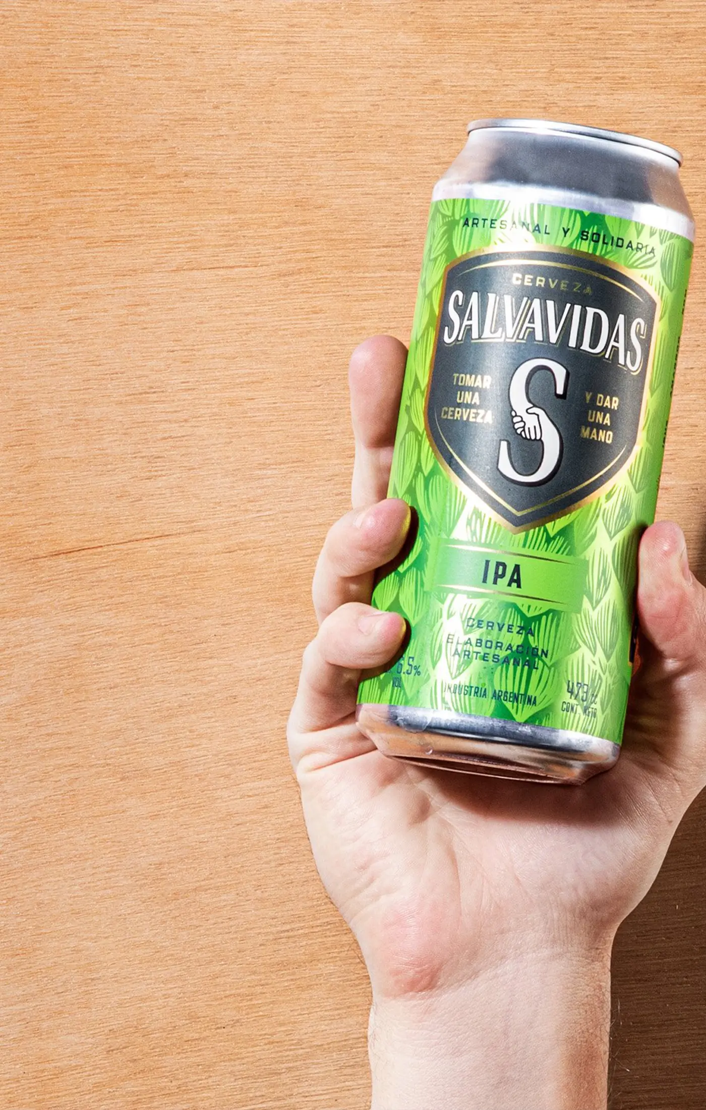 A hand holding a green can of Salvavidas IPA beer against a light wood background. The can's design is vibrant with bold branding and a refreshing tone.