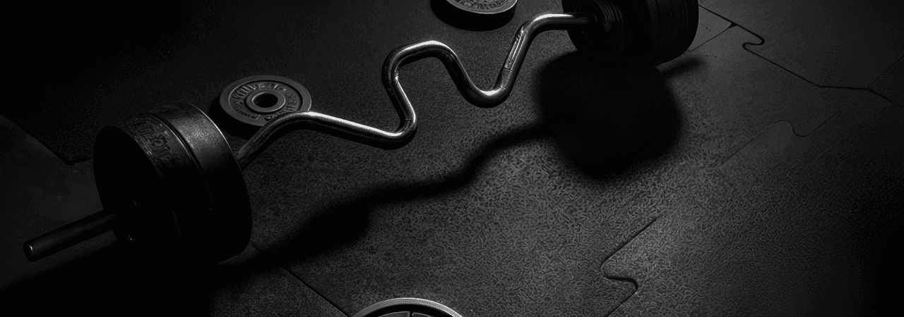 A dimly lit gym with a focus on a curl barbell with weighted plates resting on interlocking rubber floor tiles.