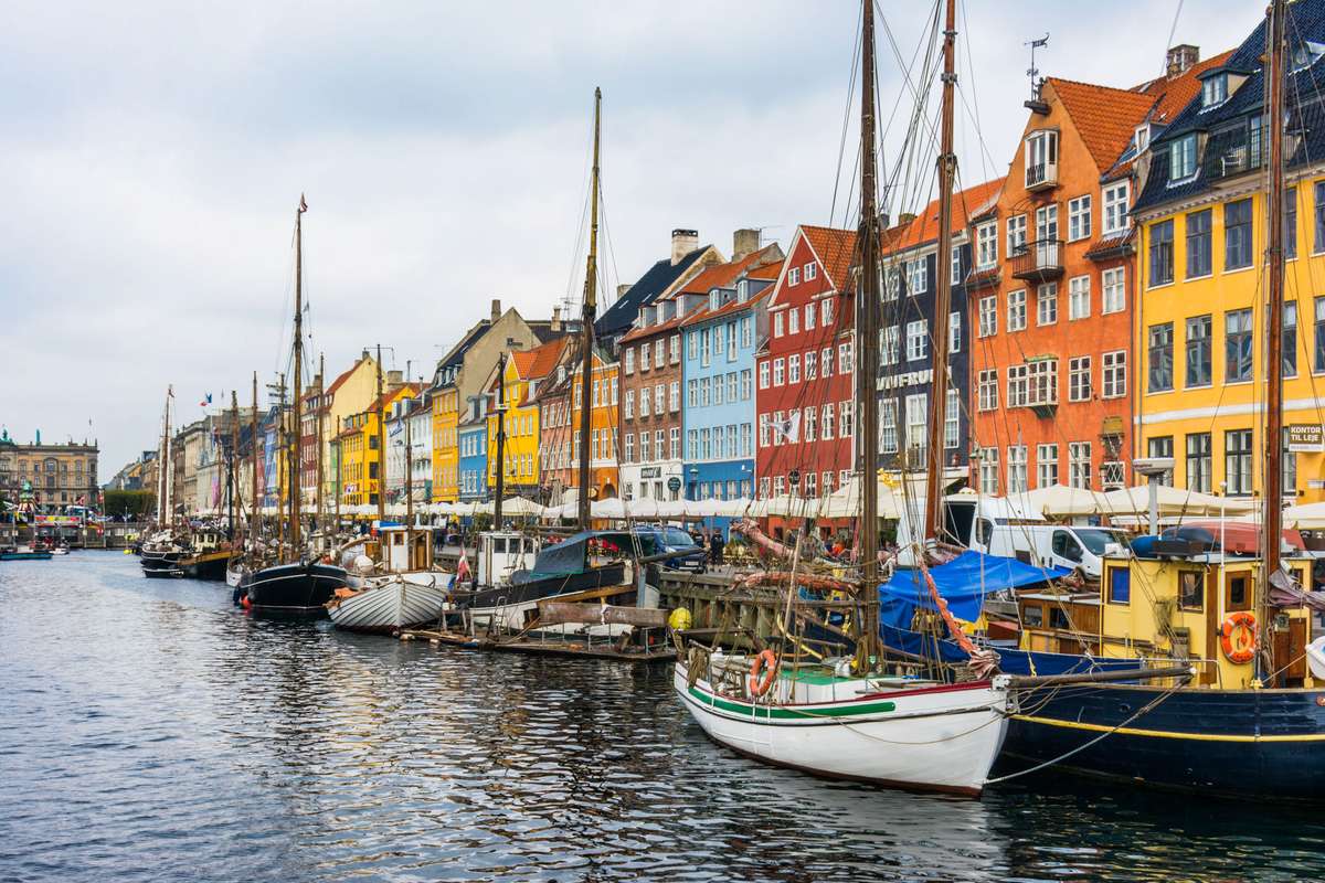Colorful multi-story buildings line a canal in Nyhavn, Copenhagen, with sailboats and small boats moored in the water.