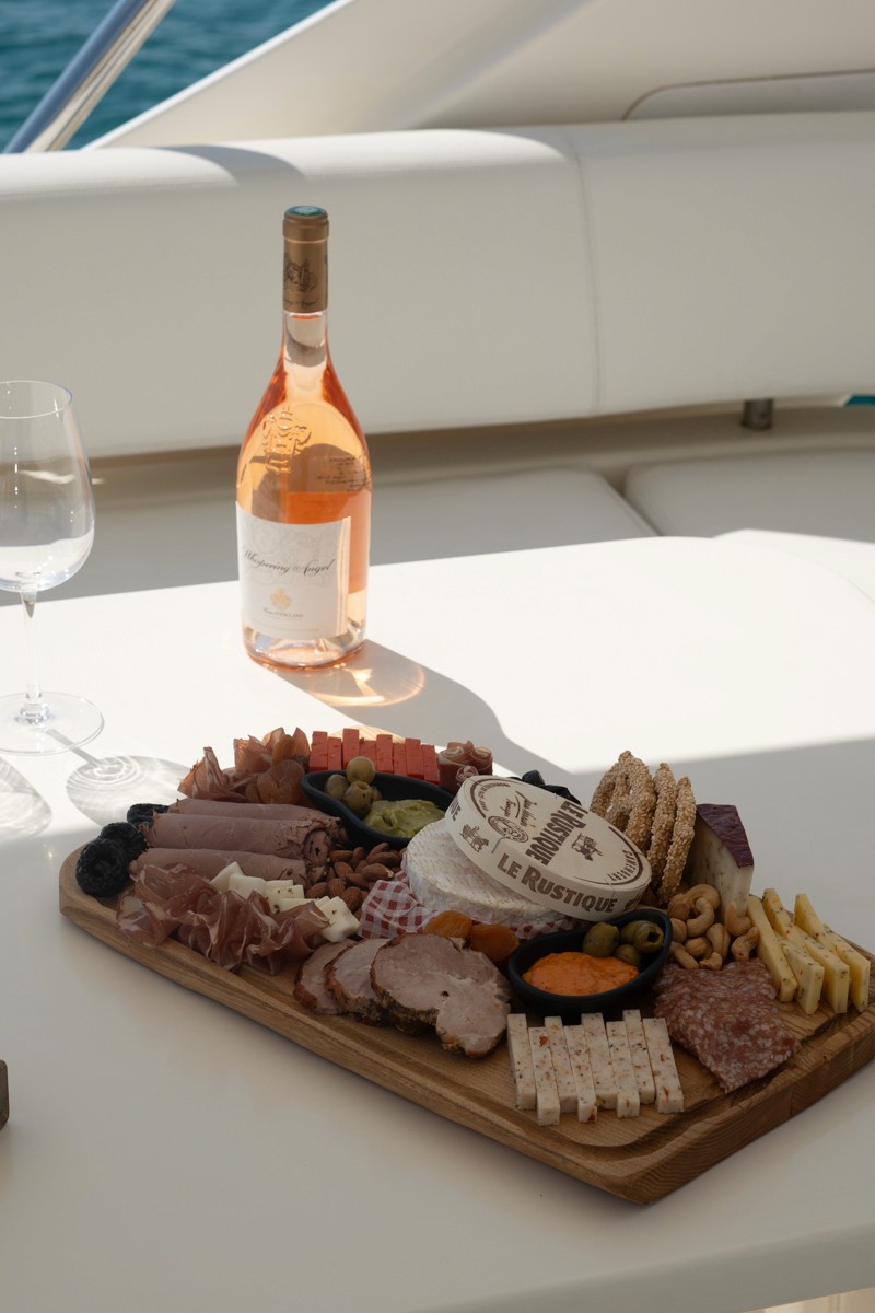 Gourmet charcuterie board with meats, cheeses, and rosé wine served on white yacht table with turquoise sea in background.
