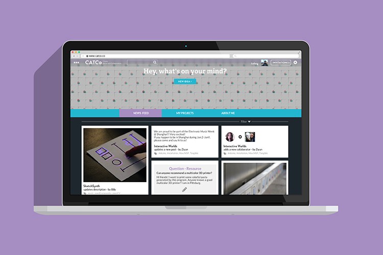 A laptop screen displays a website with a purple background. The top bar shows the website name “CATCO,” a search bar, and a navigation bar with tabs labeled “NEWS FEED,” “MY PROJECTS,” and “ABOUT ME.” Below the navigation bar, three images are shown: one of a book titled “Interactive Worlds,” another showing a multicolored 3D printer, and a third showing a wall with vertical lines. The text on the website includes “Hey, what’s on your mind?” and a “NEW IDEA” button.