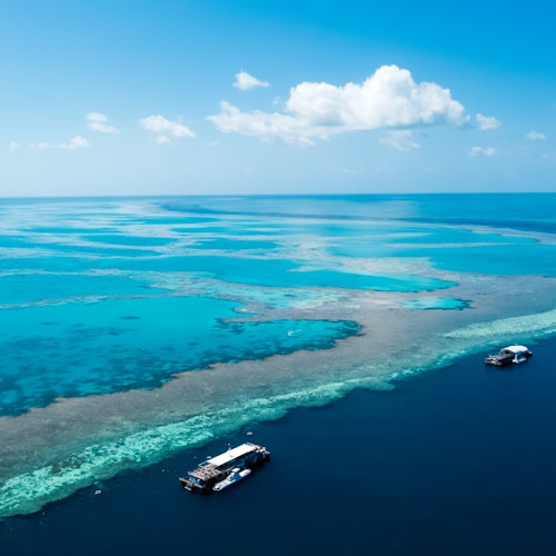 兩隻小船停泊在一個清澈的碧綠色水域的珊瑚礁附近，碧藍的天空上飄著幾朵零星的雲。
