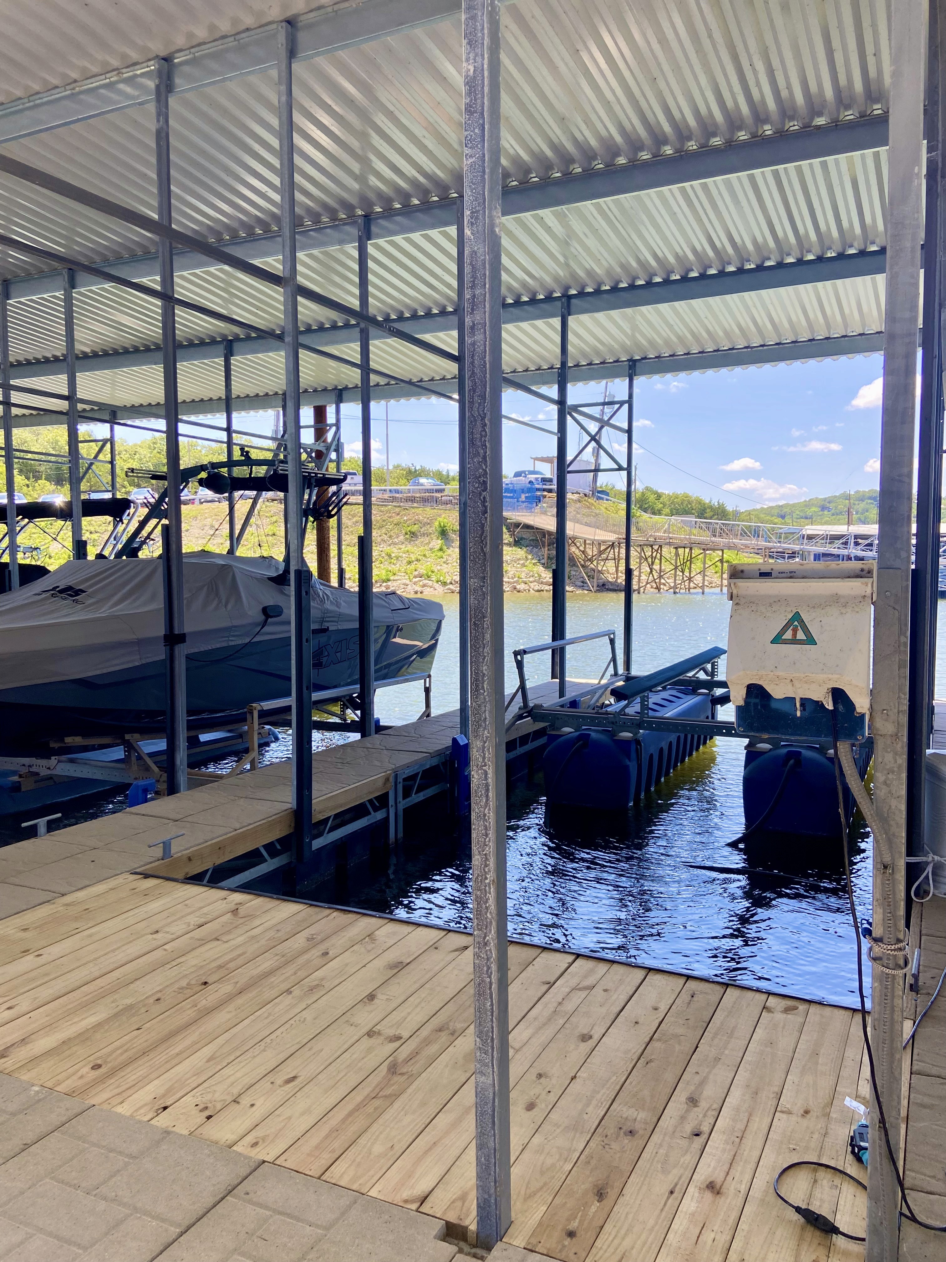 The image showcases a marina scene with several boats docked under a metal-roofed shelter, featuring blue waters and a distant view of a bridge against a partly cloudy sky.