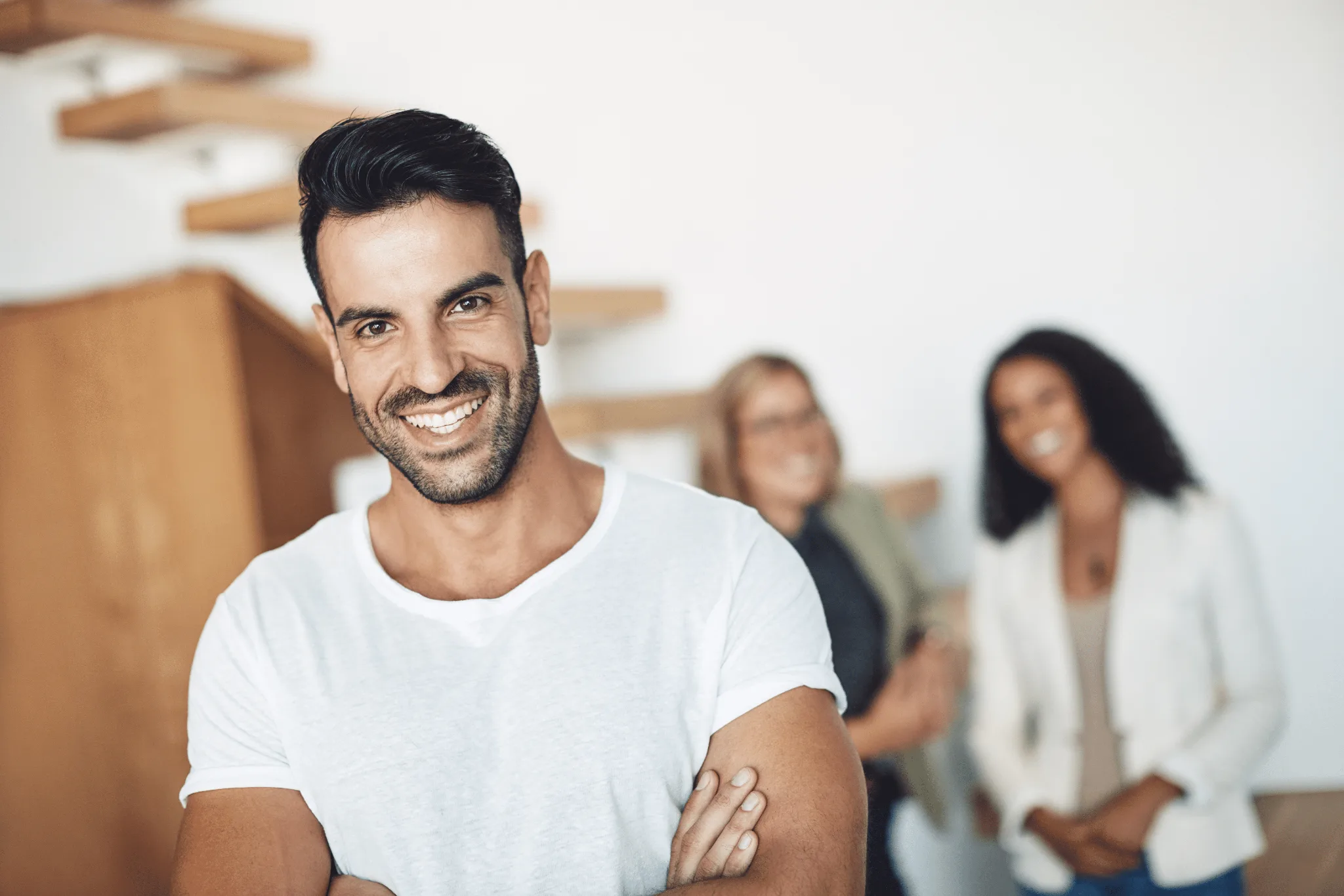 Male wearing white shirt, smiling with his arms crossed on his chest, two females blurred out at the back.