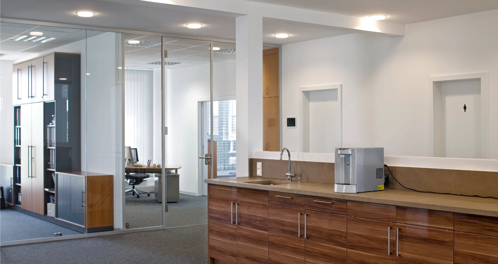 Moderne Büroküchenzeile mit Holzfronten, Spülbecken, Wasserspender und Blick in ein Büro mit Glaswänden.