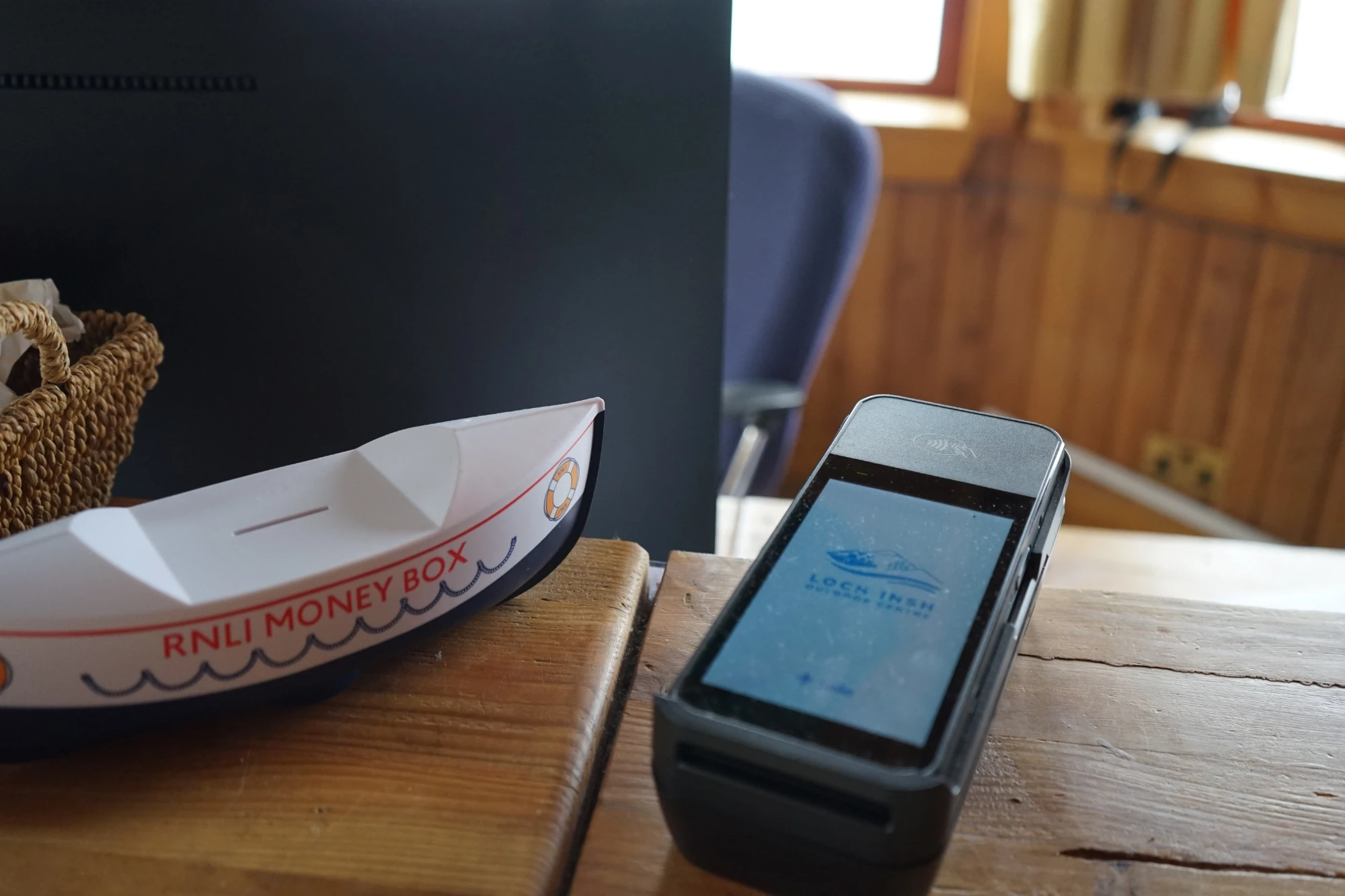 A small boat-shaped money box labeled "RNLI Money Box" is on a wooden surface beside a card payment terminal. The background shows a cozy, wood-paneled room.
