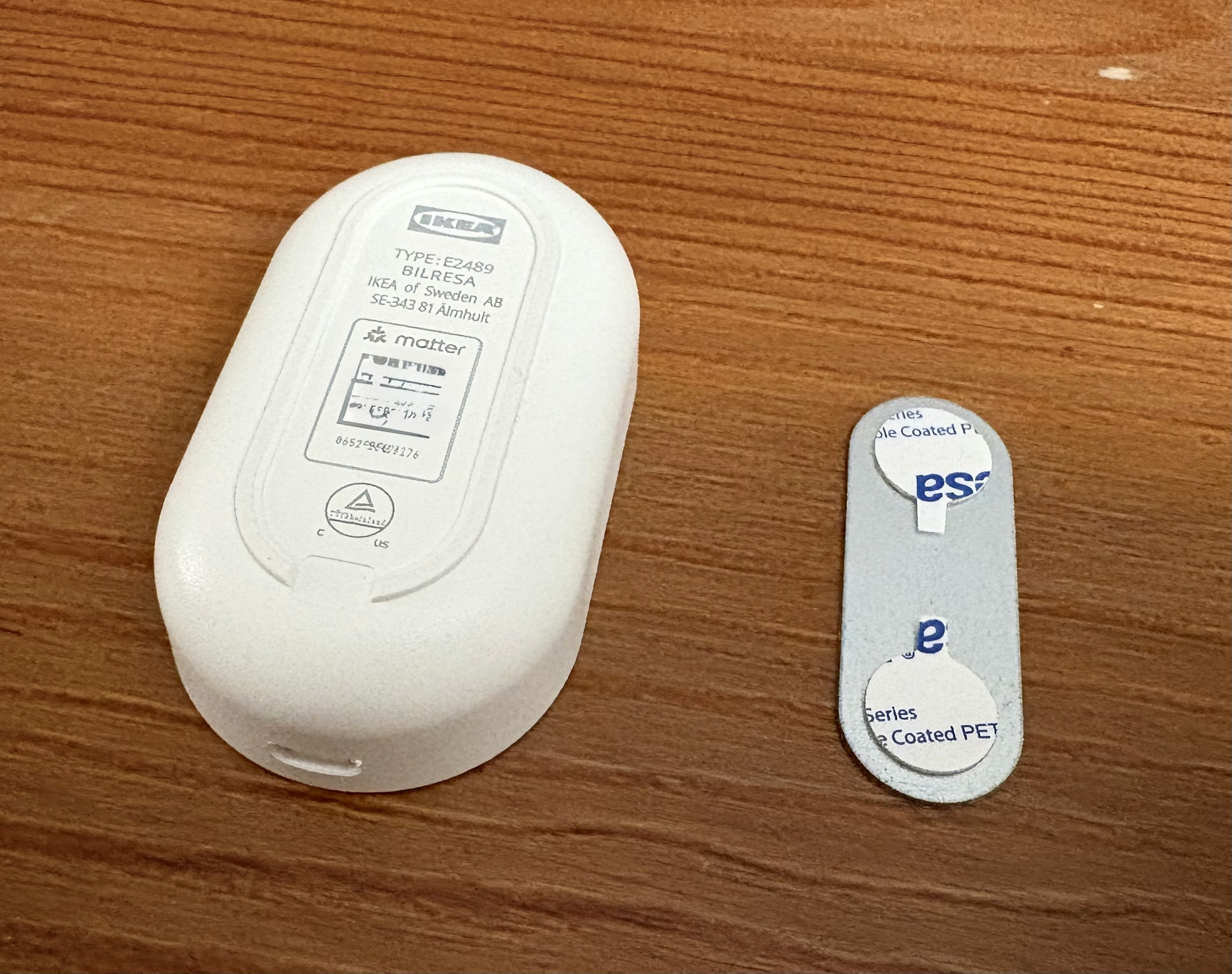 The image shows a white electronic device labeled "IKEA" alongside a small, flat metallic strip with markings, all placed on a wooden surface, possibly related to a household system for energy management or smart home integration.