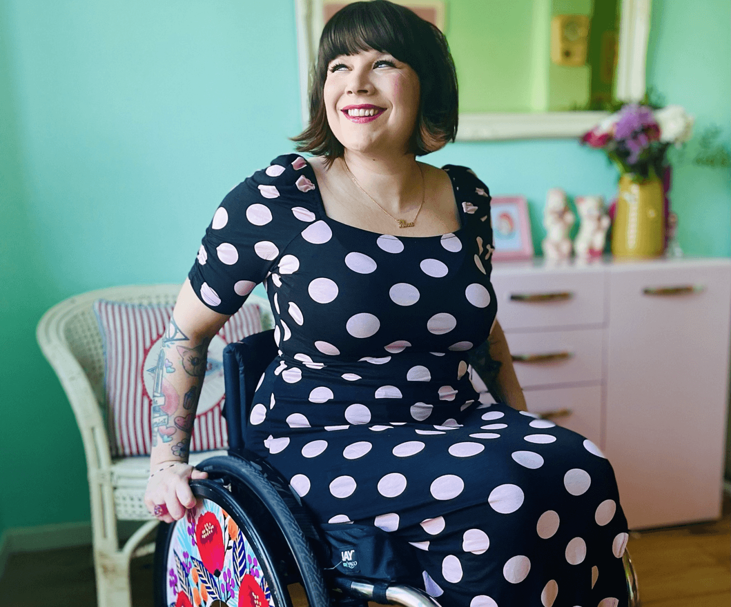 Nina is sitting in her wheelchair with flower print wheel cover, Ewearing a long navy blue fitted dress with white polka dots, with a big smile on her face.