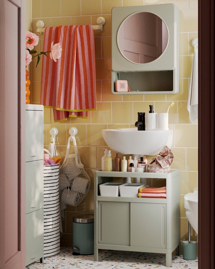 A modern bathroom featuring pastel-colored tiles, a round mirror, a wall-mounted cabinet, a circular sink on a green storage unit, neatly organized toiletries, and striped towels hanging prominently.
