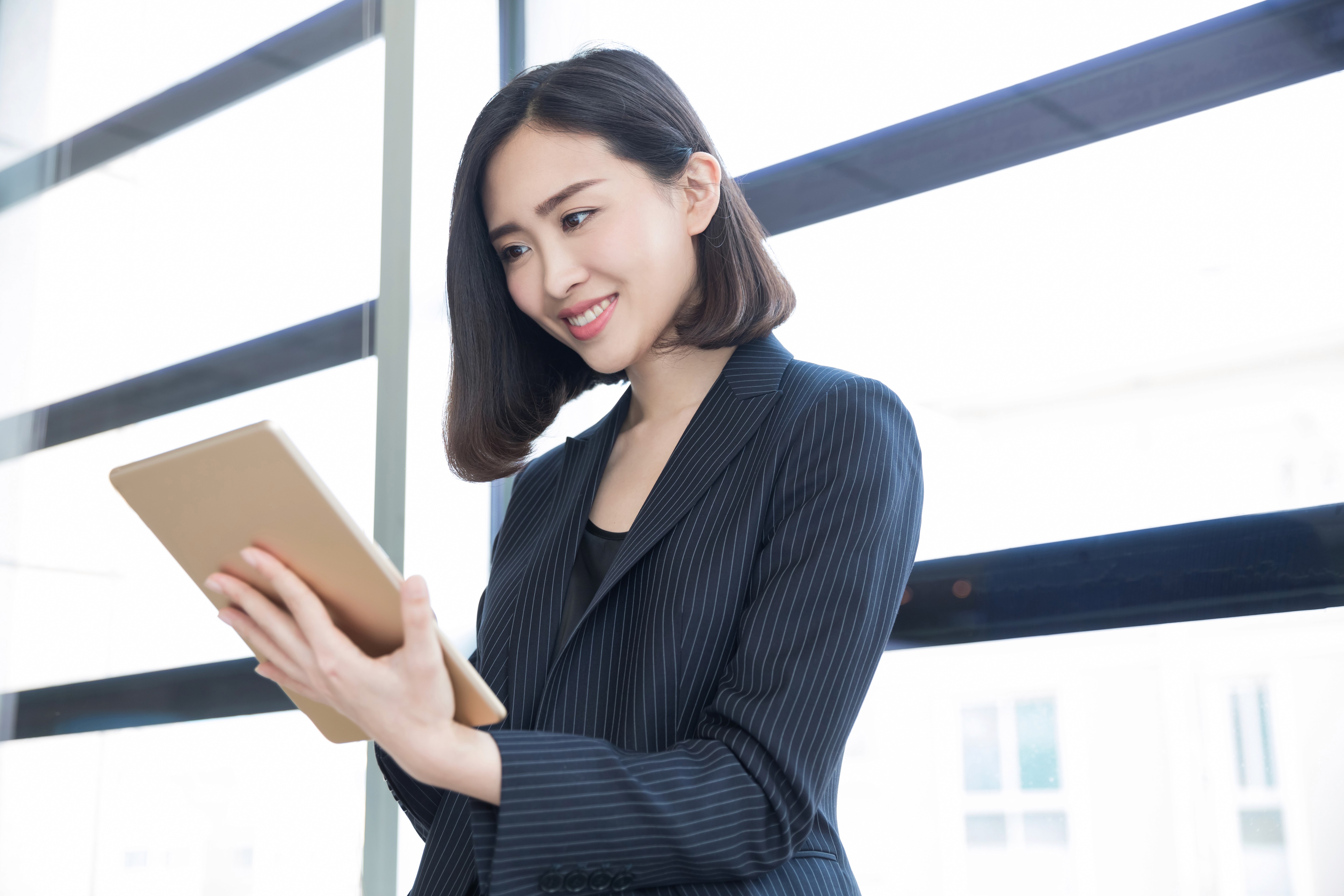 A smiling woman in business attire holds a digital tablet while standing near a window, representing real-time monitoring and supervisory tasks.