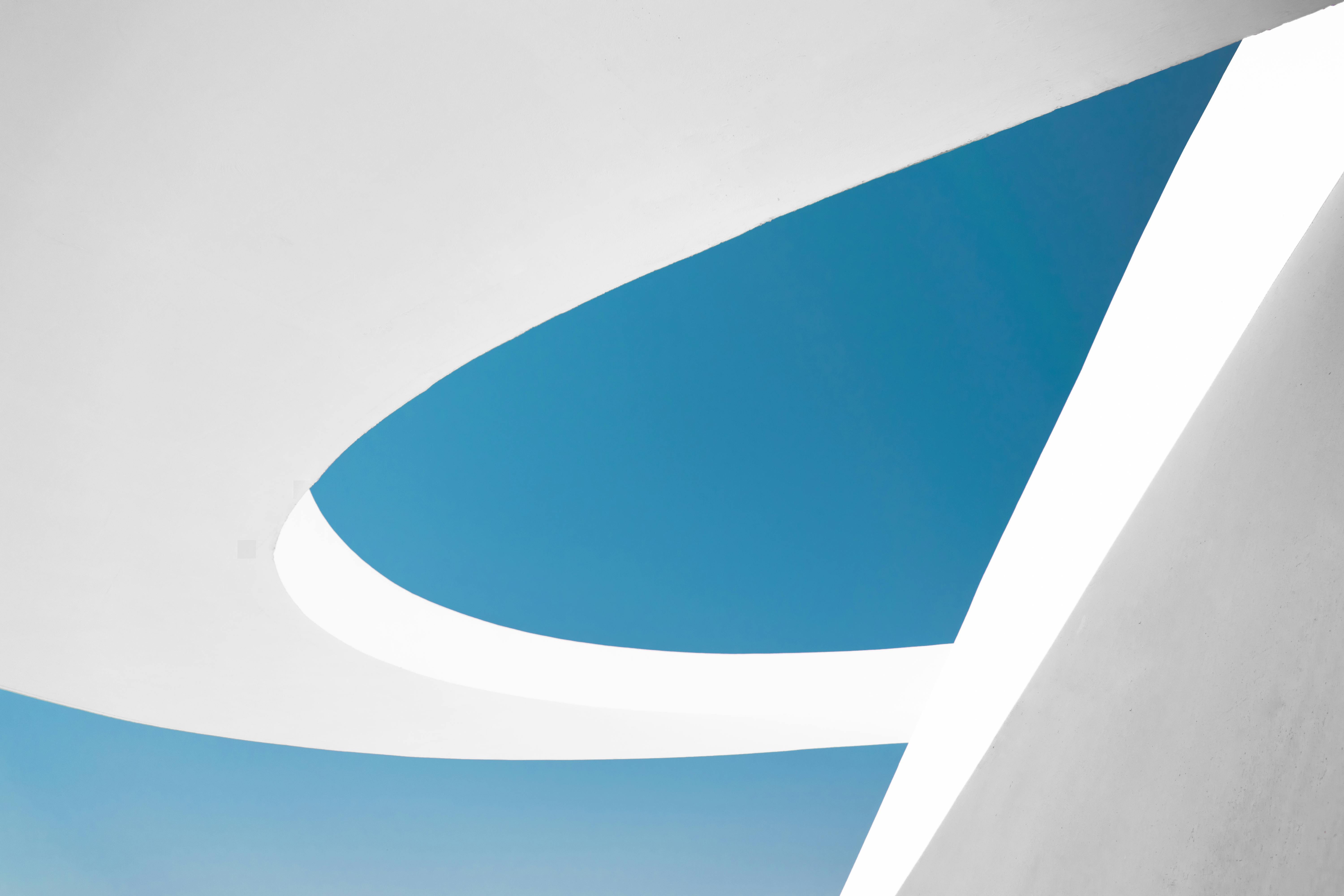 A minimalist architectural scene featuring smooth white curved structures framing a clear blue sky, with clean lines and soft shadows creating an abstract, modern composition.