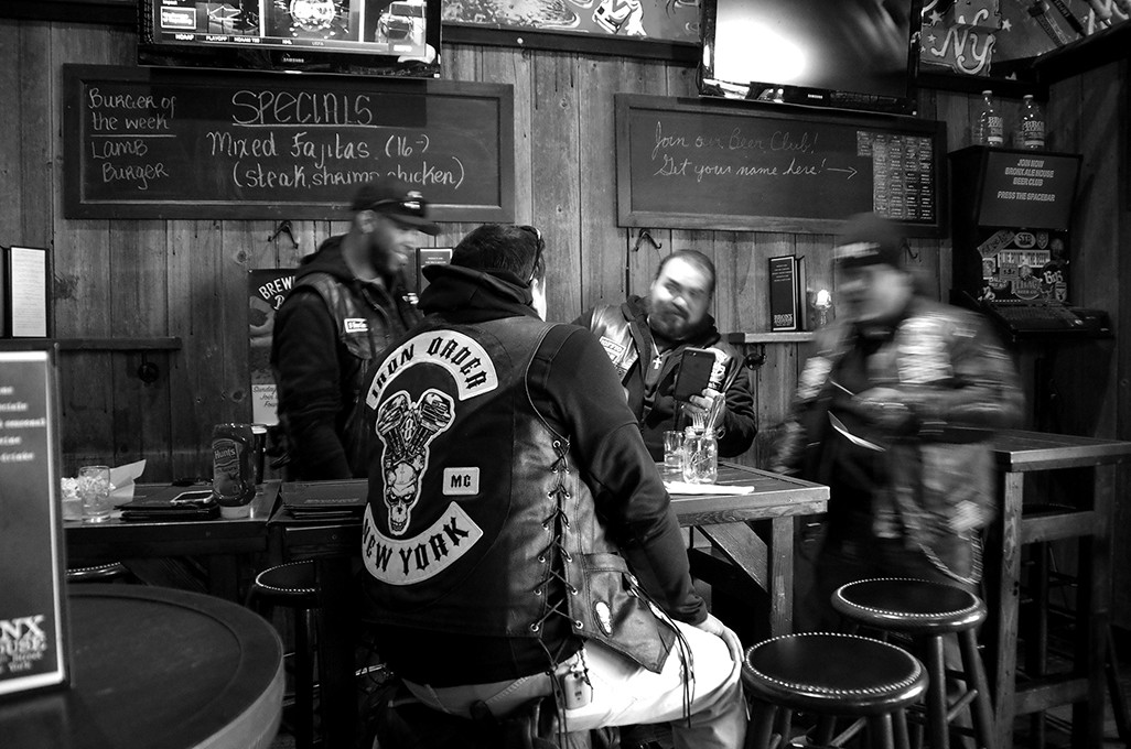 A black and white photo shows three men in a bar or restaurant. The man on the left wears a black jacket with a white skull and crossbones patch reading "THON ORDER NEW YORK." The man in the center wears a black jacket with a white skull and crossbones patch. The man on the right wears a patterned jacket and a cap. Behind them are chalkboards listing menu items including "Burger of the week," "Lamb Burger," "Mixed Fajitas," and "Steak shawarma chicken." Bar stools and tables are visible in the background.