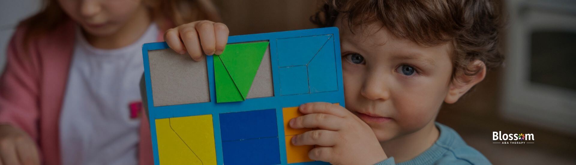 A boy with autism holds a colorful puzzle board during ABA therapy, with a girl working nearby.
