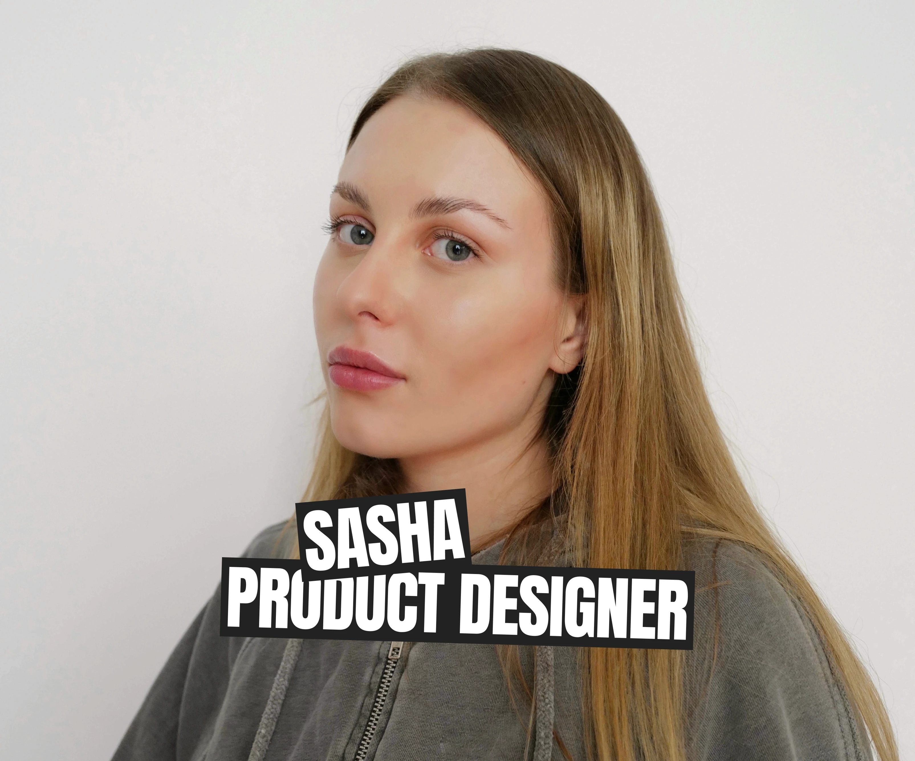  A portrait of Sasha, a product designer, against a plain white background. Sasha is wearing a gray hoodie and looking directly at the camera with a confident expression. Bold text in white over a black background at the bottom reads "SASHA PRODUCT DESIGNER." The minimalist design highlights her role, giving the image a clean and professional feel.