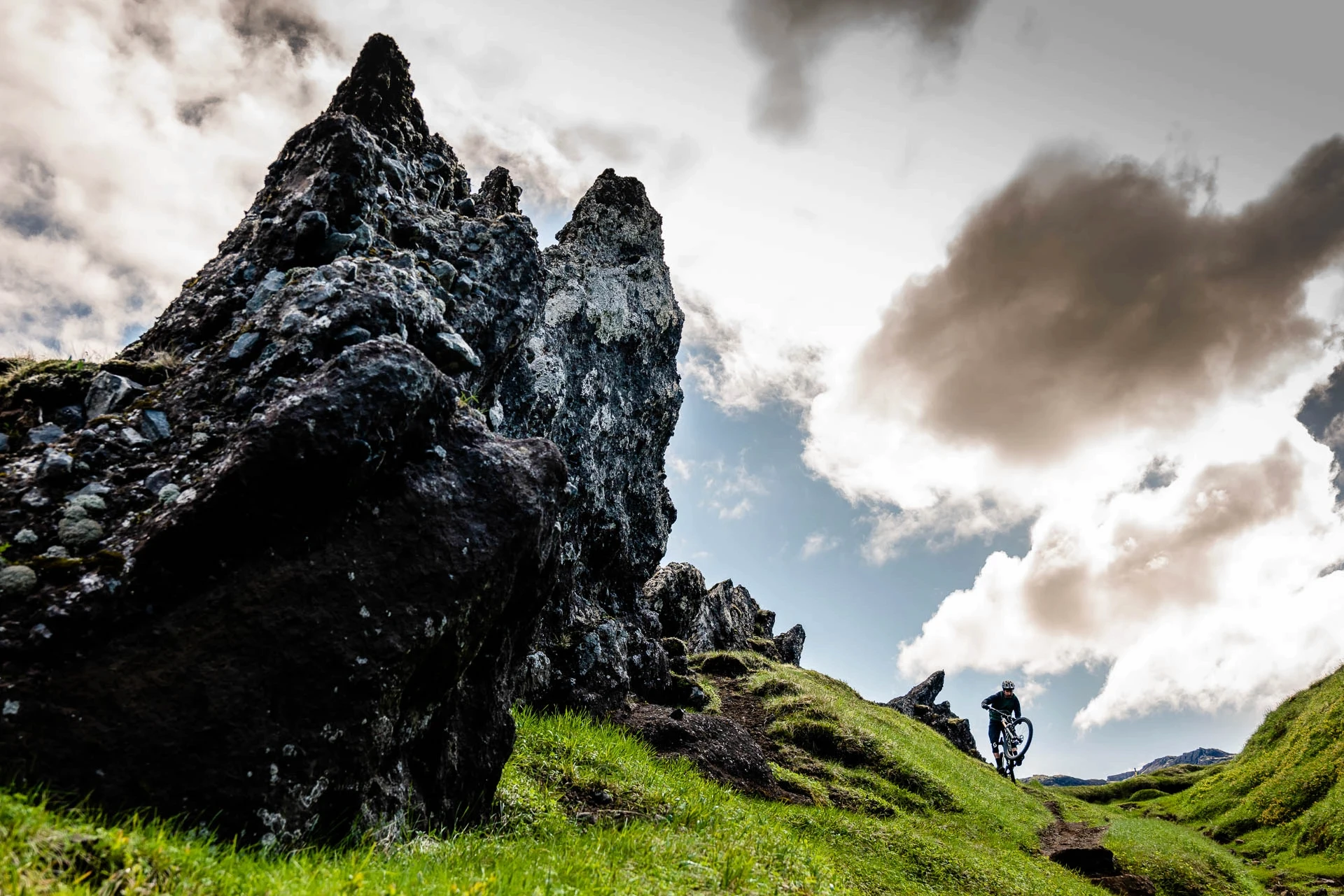Mountain biker riding along a trail beside a jagged rock spire under dramatic clouds.