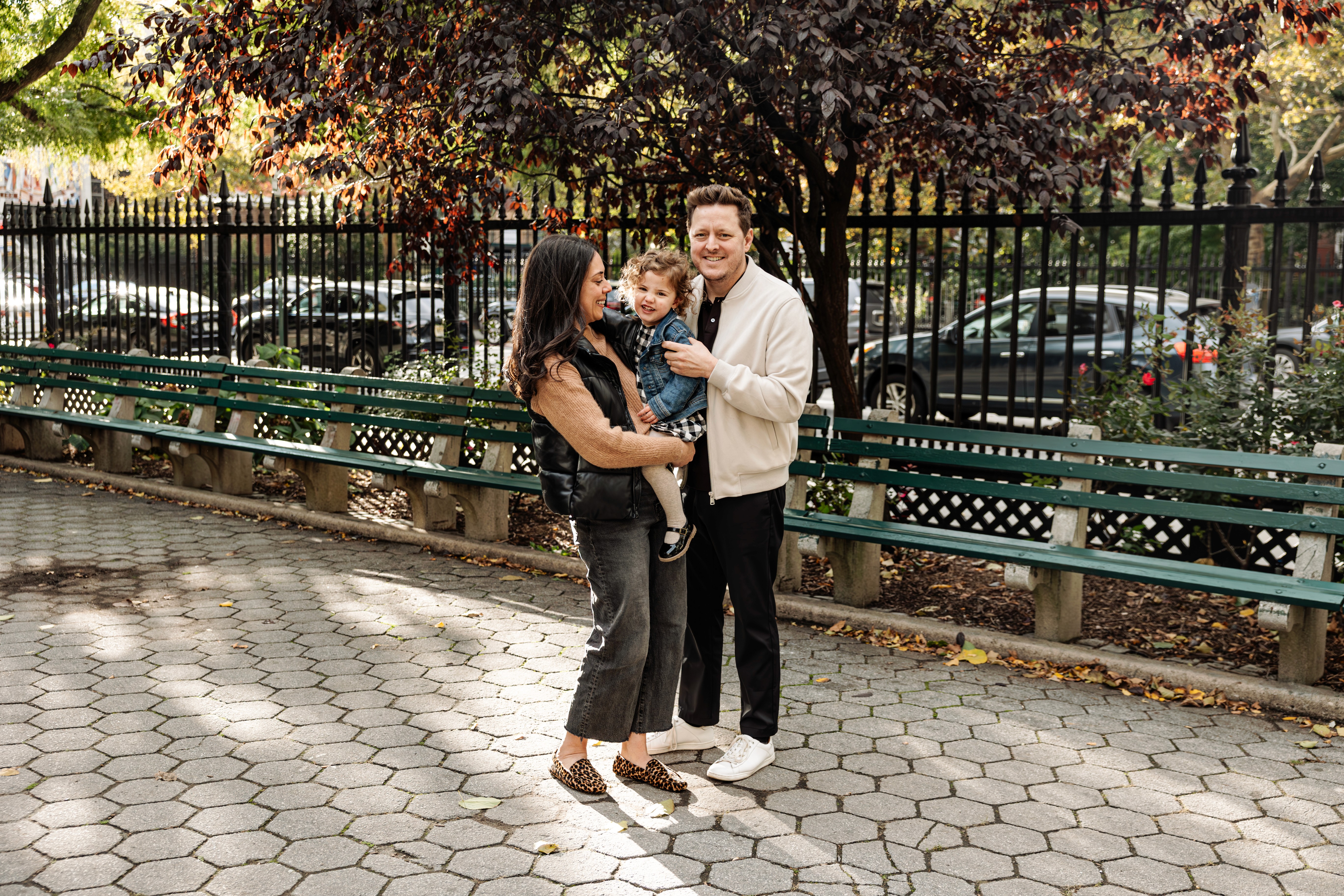 Family of three on a park bench in Stuyvesant Square, NYC, daughter nestled in the middle as all three share a warm, loving look at each other — natural, candid family photography by Lizz Spano Photography, New York City family photographer.