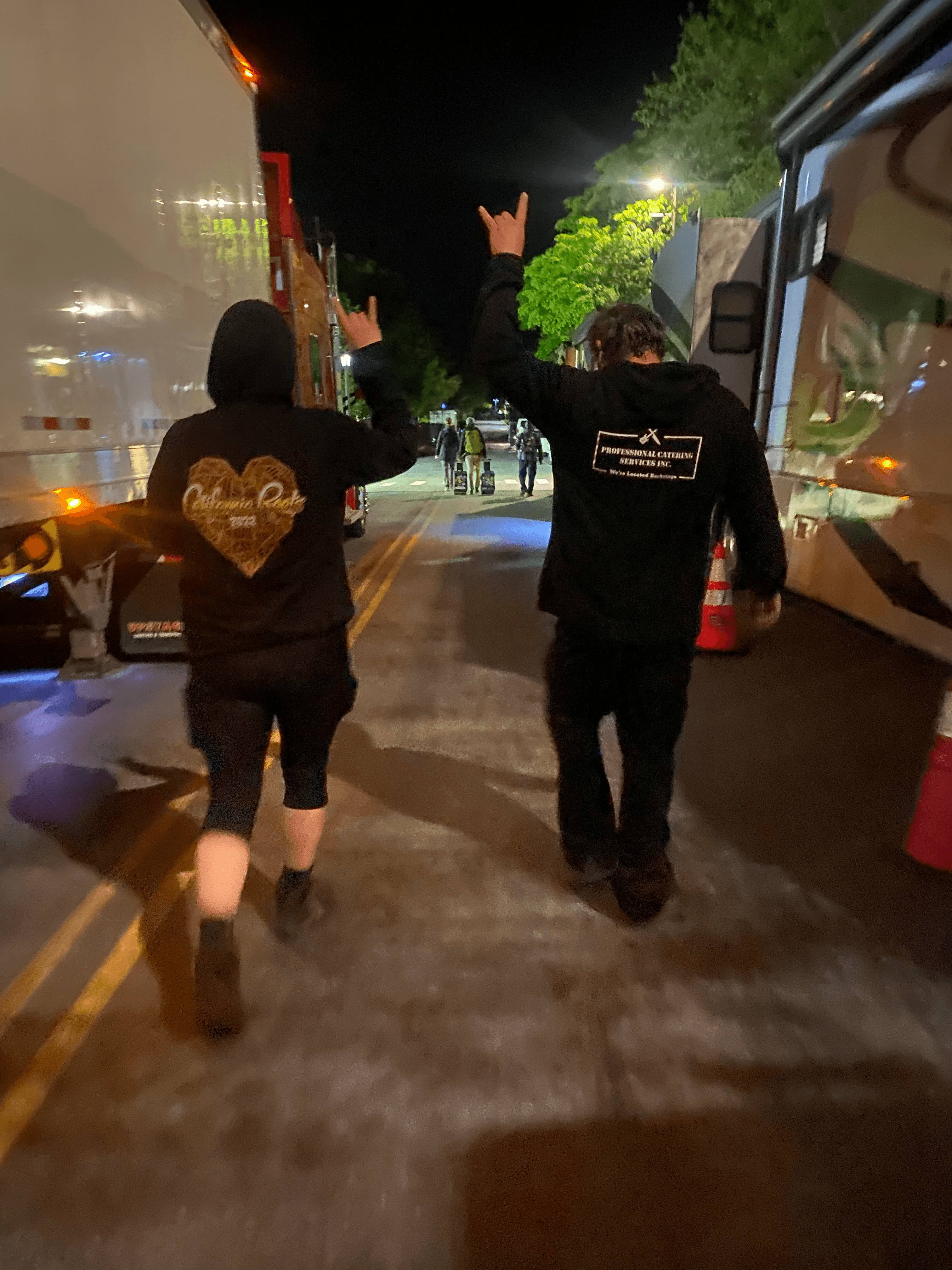 Two individuals walk hand-in-hand down a lit street at night, dressed in black hoodies.