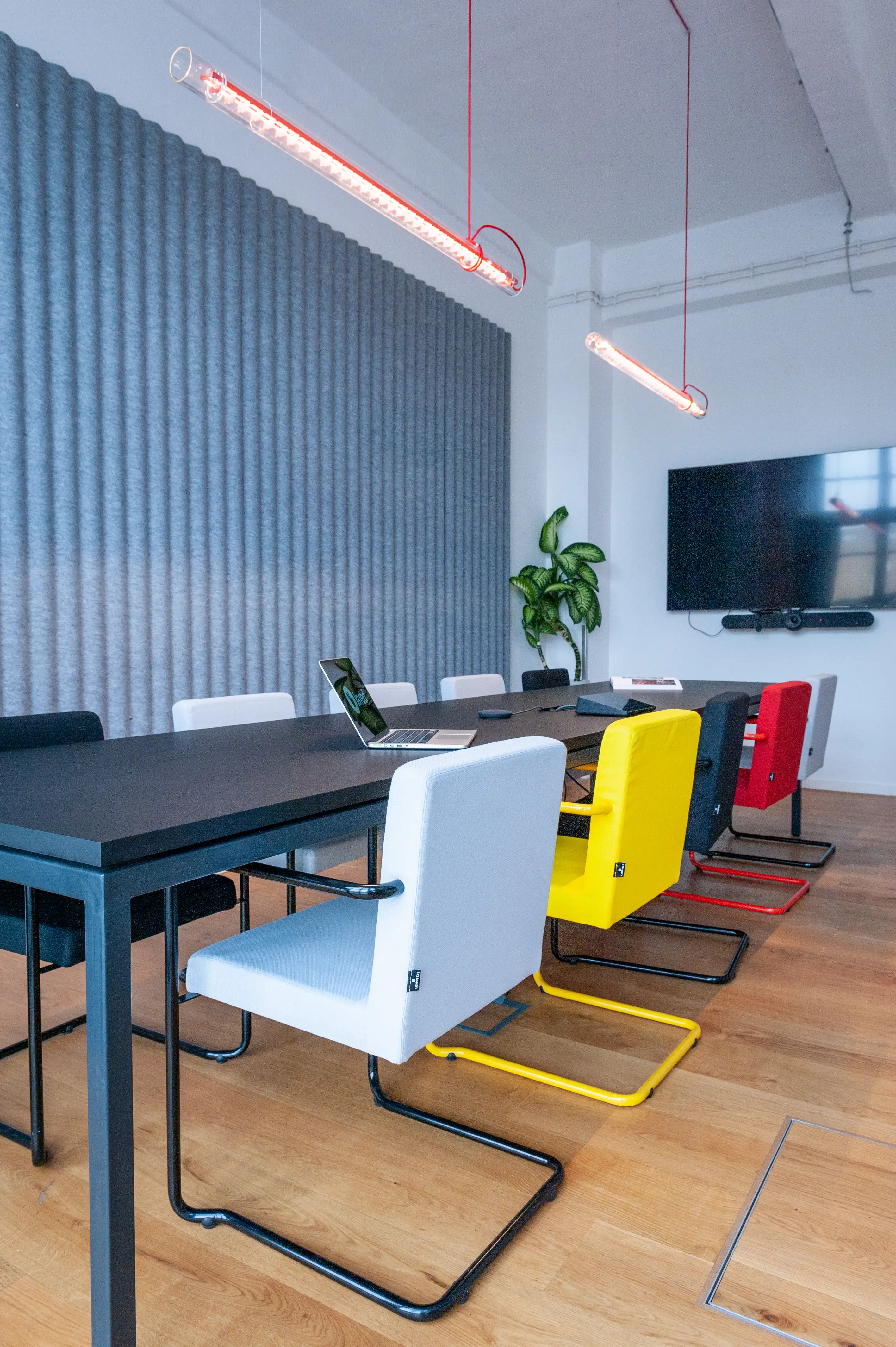 Conference room at LA RED with a long black table, cantilever chairs in white, yellow, red and black, a grey corrugated acoustic wall, red-framed tube pendant lights and a wall-mounted TV.