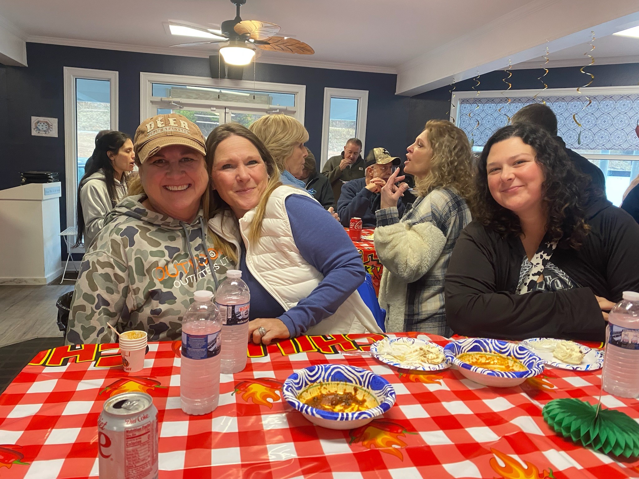 A group of people enjoying a meal at a festive gathering, seated around a table with a red and white checkered tablecloth, bowls of snacks, and drinks, in a warmly lit room with a relaxed atmosphere.