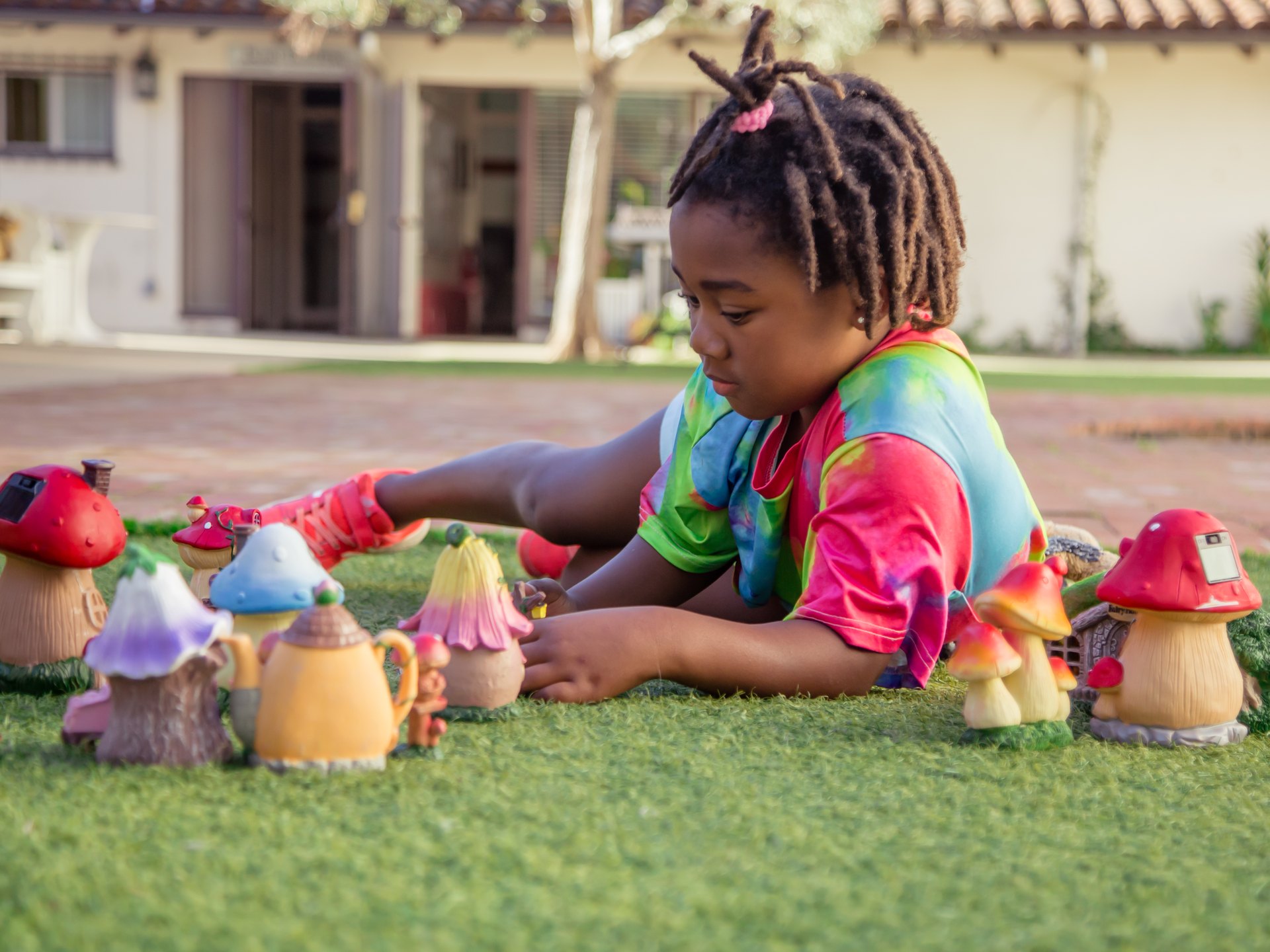 Student in a tie-dye shirt lying on the grass playing with miniature fairy-garden mushroom figures