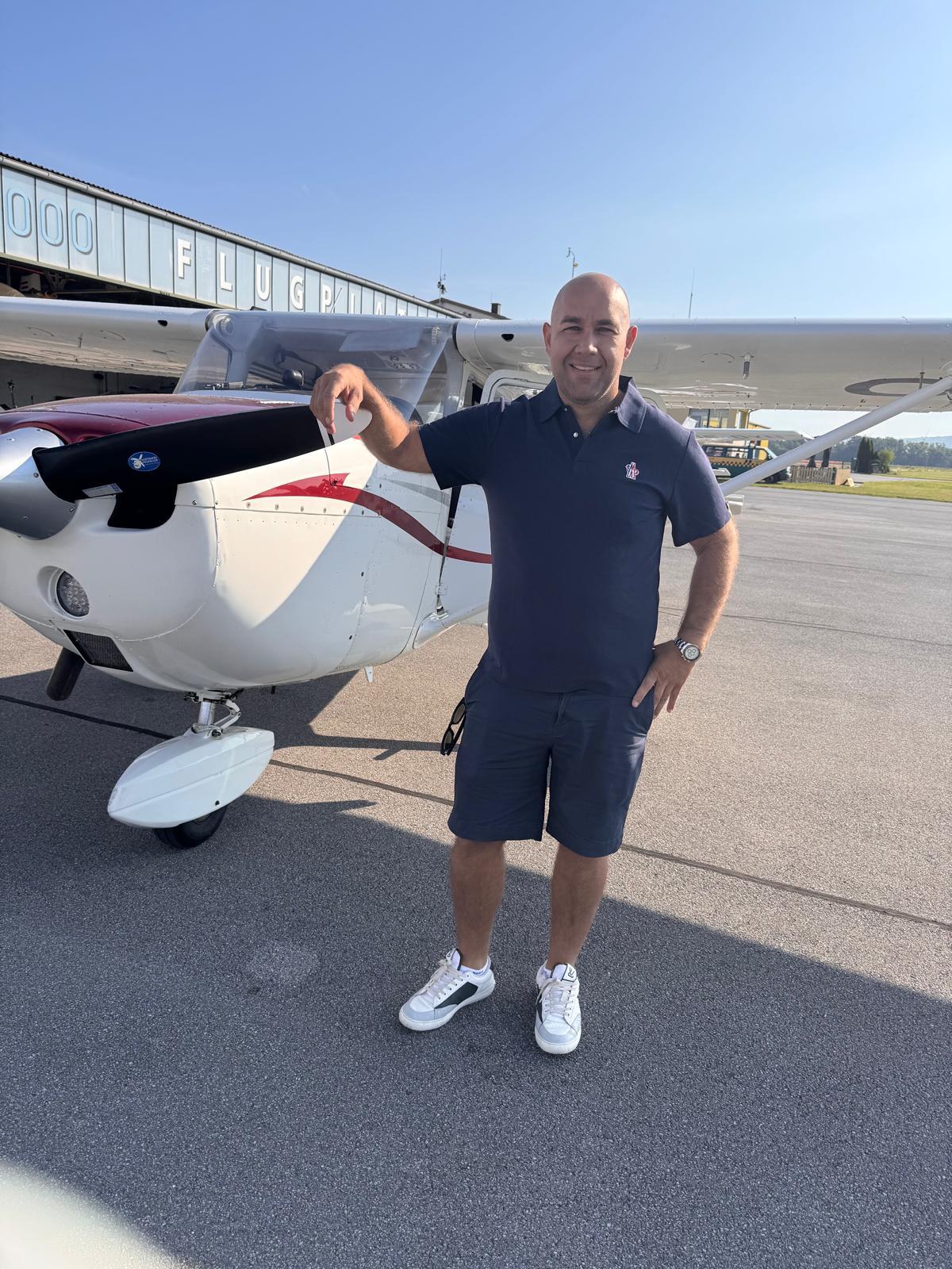 A man in a light-colored suit and sunglasses relaxes on a boat, with mountains and water in the background.