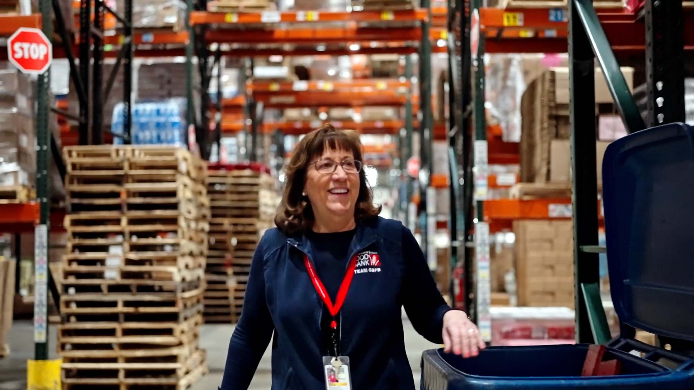 Woman wearing glasses and a navy “Food Bank” vest with a red lanyard stands inside a warehouse aisle with stacked pallets and shelving behind her.