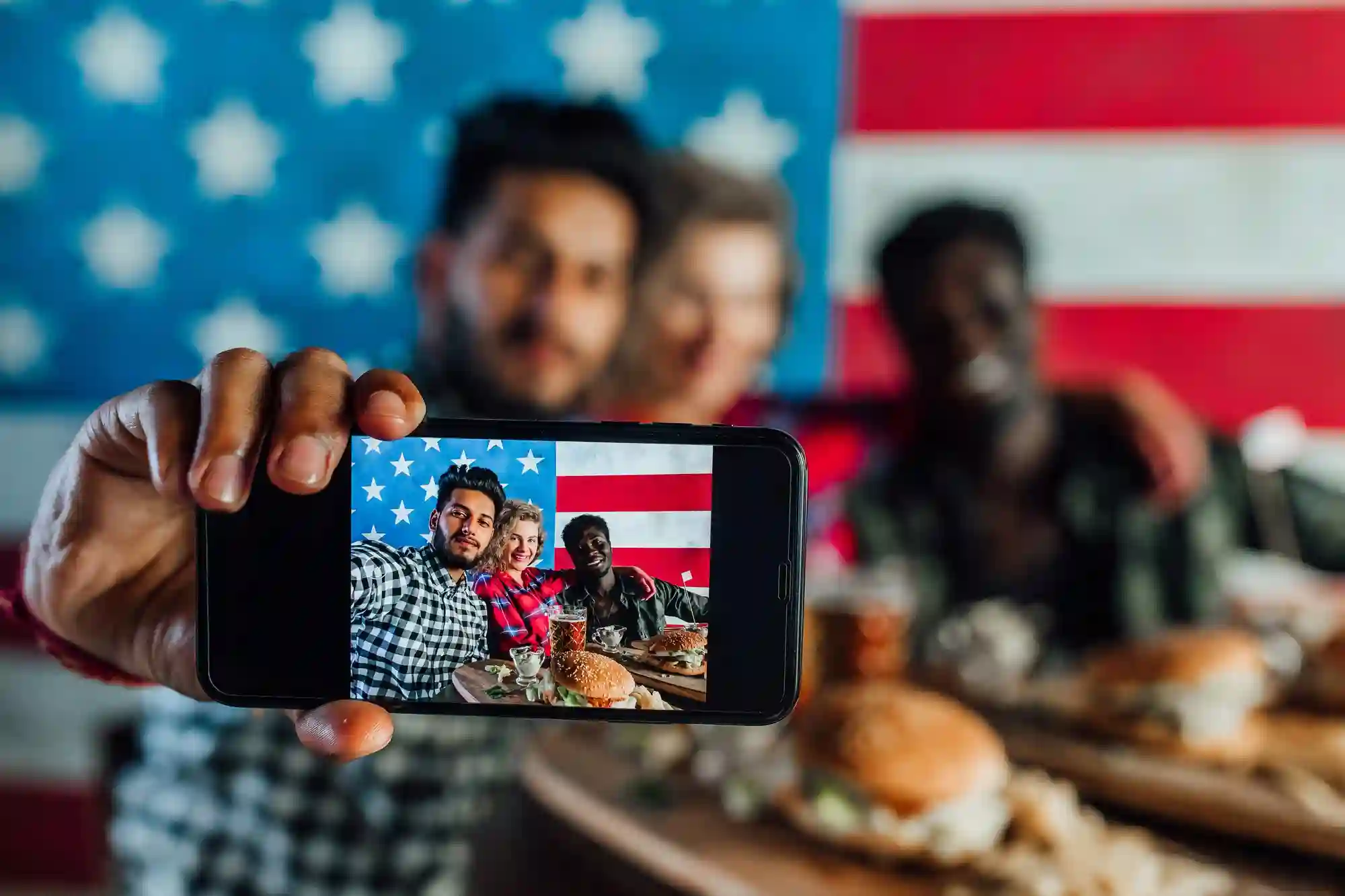 A diverse group of friends taking a selfie at an American-themed dinner with burgers and a large flag.