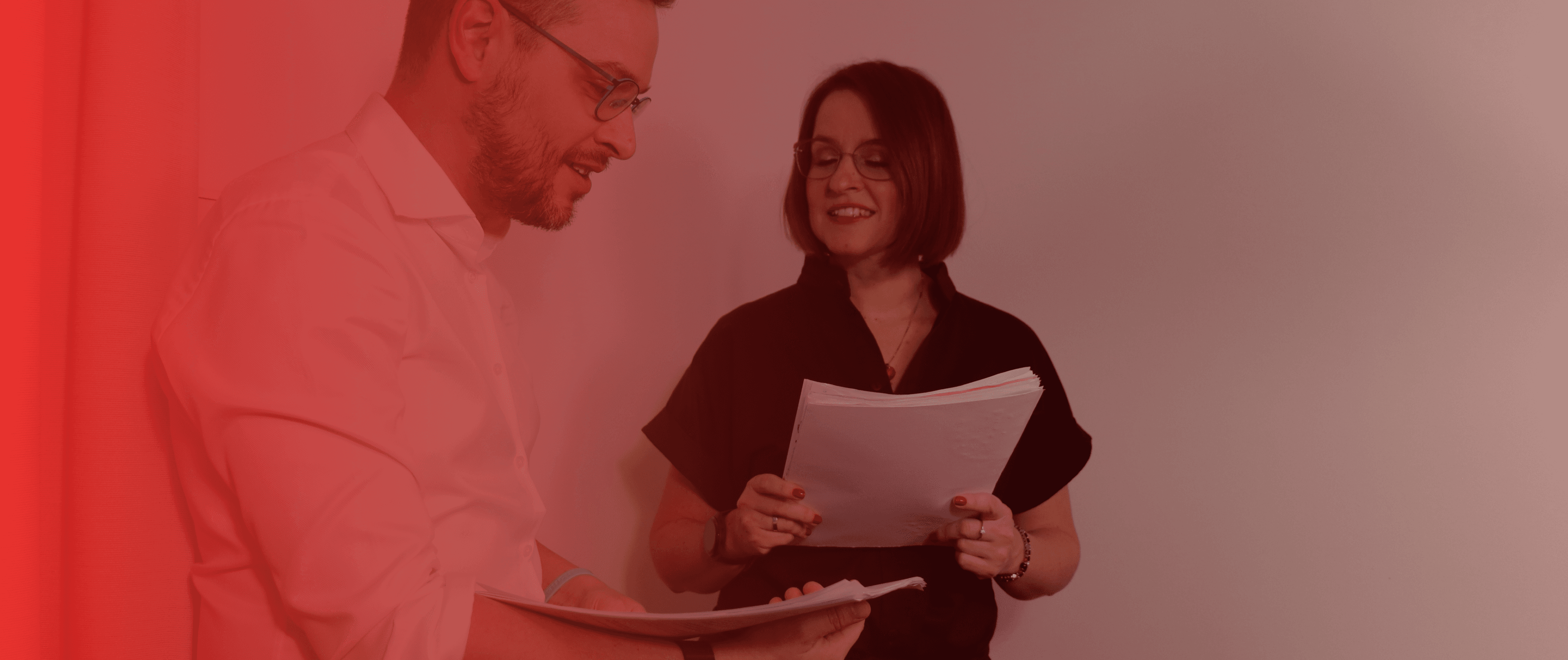Muž a žena diskutují o dokumentech, stojící před červeným pozadím. Oba vypadají, že jsou zabráni do rozhovoru.