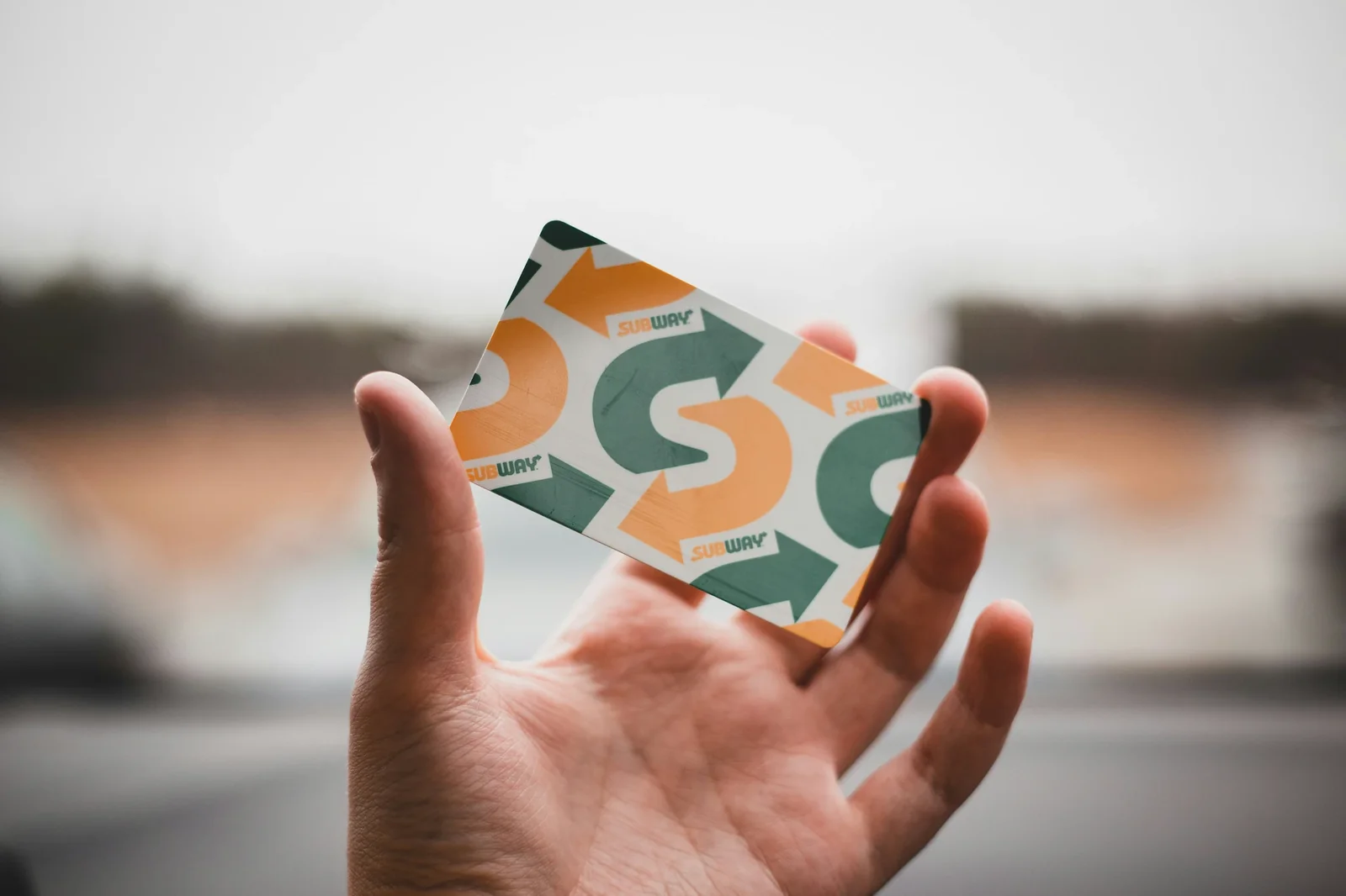 Hand holding Subway loyalty program card with orange and green arrow pattern against blurred urban background.