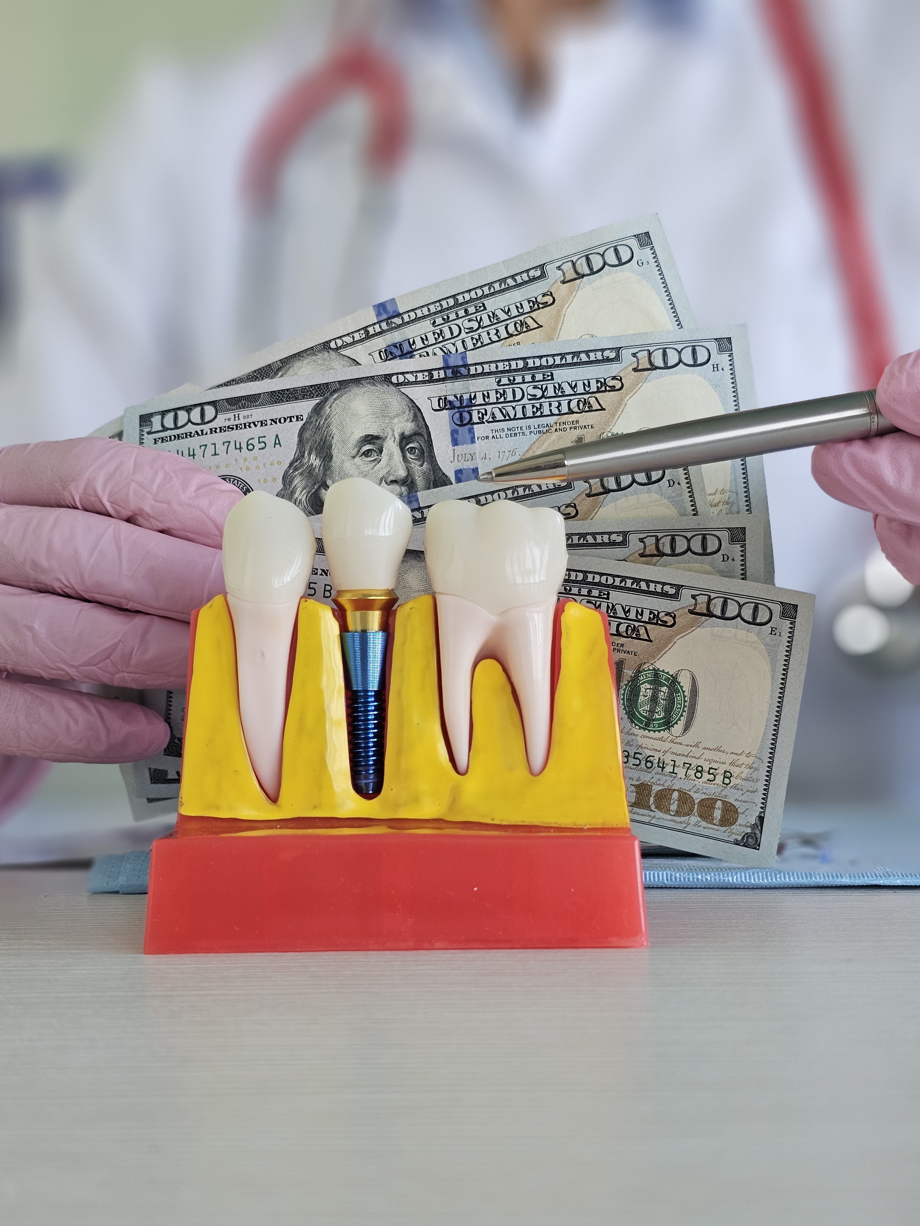 A dentist wearing pink gloves holds dental tools and a model of teeth with a dental implant, while U.S. hundred-dollar bills are visible in the background, highlighting the financial aspects of dental care. 